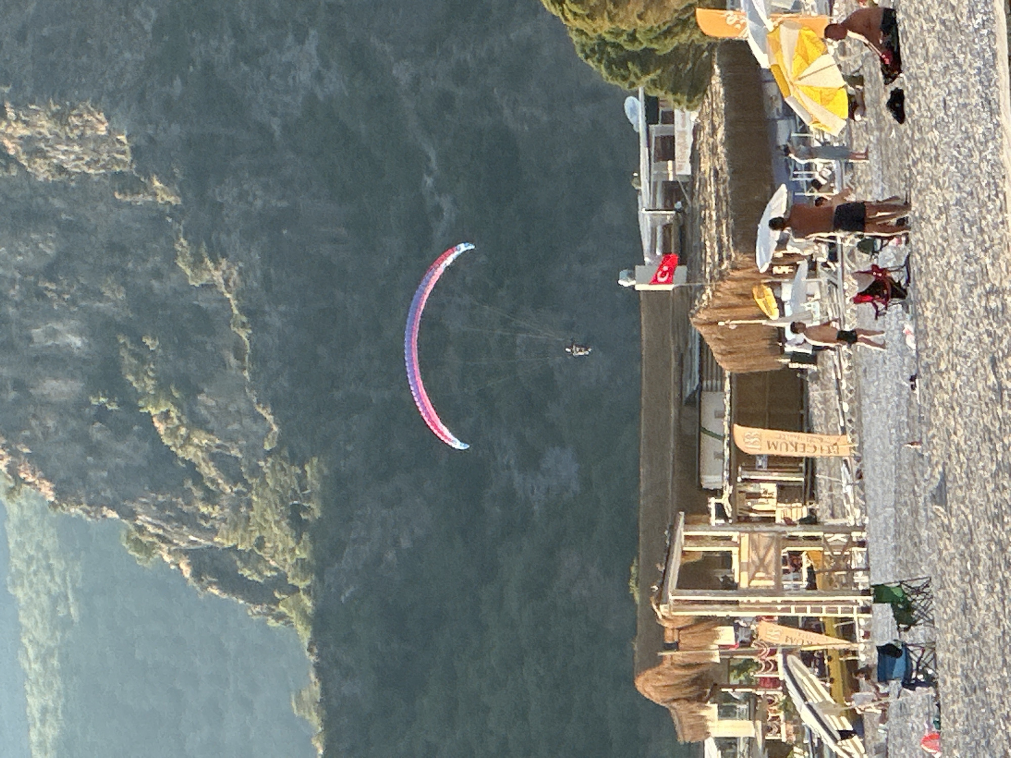 The image captures a vibrant coastal scene in Fethiye, Turkey, likely near Ölüdeniz. The view is from an elevated perspective, looking down towards a beach resort, the sea, and forested mountains.
In the upper-left, a large, verdant mountain slopes down towards the deep blue-green water. A colorful paraglider, striped in pink, blue, and white, is prominently featured in the mid-air above the water, carrying a single person. Below the paraglider, another, smaller paragliding tandem can be faintly seen.
To the right, a sandy beach runs horizontally, densely populated with beachgoers and resort infrastructure. Numerous people are visible on the sand, some standing, some seated on sun loungers or in beach chairs under various sun umbrellas (yellow, white, and thatched). Buildings and structures of a beach club or hotel line the back of the beach, featuring wooden accents and thatched roofs. A Turkish flag, red with a white star and crescent, is displayed on one of the buildings.
The time of day appears to be late afternoon, with warm, directional sunlight creating soft shadows. The sky is clear, suggesting pleasant weather. Visible text on flags includes "Biçerim" and "MAVİ KÖŞE" (meaning "Blue Corner" in Turkish), likely identifying beach establishments. The overall atmosphere is one of leisure and outdoor activity in a scenic coastal setting.