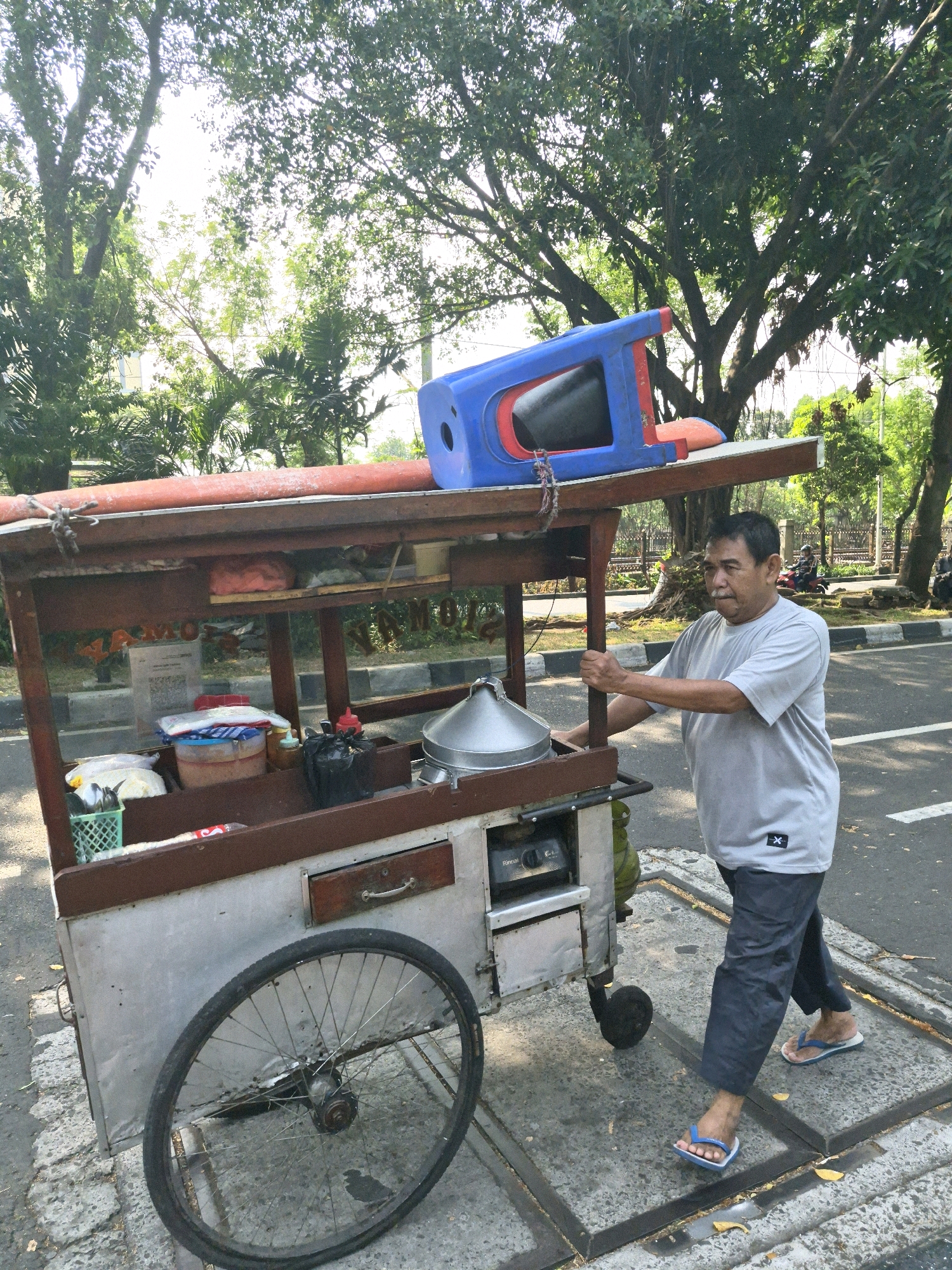 A middle-aged man with a mustache and short, dark hair is pushing a food cart on a paved surface next to a street. He is wearing a light gray t-shirt with a black logo on the left chest and dark blue pants. He has on blue and white flip-flops. The food cart is made of metal and wood and has a large silver steaming pot on top, with various containers and items stored within its shelves and compartments. A blue and red plastic item is mounted on top of the cart. The background shows a tree-lined street with dappled sunlight. The man is looking to his left and is in the process of moving the cart. The scene appears to be outdoors in an urban environment, likely during the daytime.