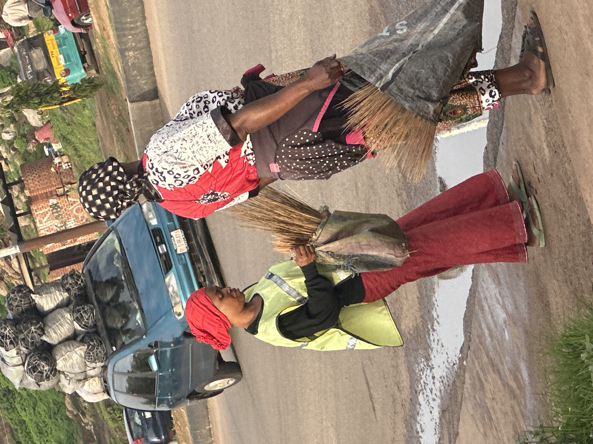 The image depicts a street scene in Gura Top, Nigeria. Two women are walking on a paved road. The woman in the foreground, on the left, is wearing a red headscarf, a bright yellow vest with reflective strips over a black long-sleeved shirt, and a red skirt. She is carrying a bundle of brooms in a sack over her right shoulder. The woman behind her, on the right, is wearing a black and white checkered headscarf and a brightly colored top with red, white, and black patterns. She is carrying a large, dark-colored sack that appears to contain brooms as well. In the background, a teal minivan is parked on the side of the road, with a large load of sacks on its roof. A red car and a three-wheeled vehicle are also visible further down the road. The environment appears to be urban or peri-urban, with some vegetation and brick structures in the background.