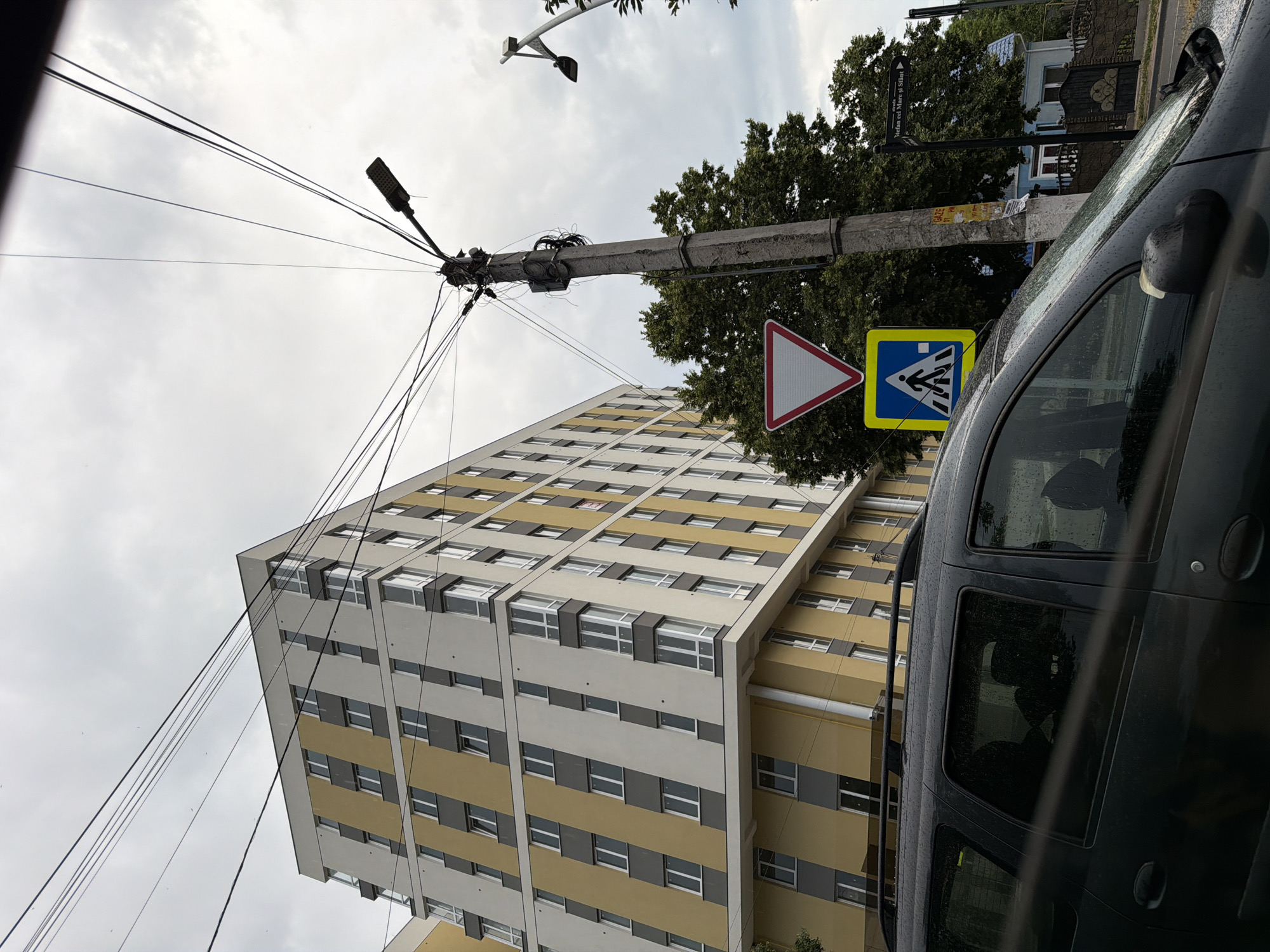The image shows a street scene in Strășeni, Moldova, under an overcast sky. A modern, multi-story building with yellow and white facades dominates the left side of the frame. In the foreground, the side of a dark-colored vehicle, possibly a station wagon, is visible. The vehicle's windows are wet, indicating recent rain. A utility pole stands to the right of the building, cluttered with numerous wires and a street lamp. Two traffic signs are attached to the pole: a triangular yield sign with a red border and a square sign with a blue background depicting a pedestrian crossing. Behind the utility pole, a large tree with dense green foliage is present. Further in the background, a small, light-blue house with a white trim and a dark gate can be seen. A street sign with Romanian text, "Stefan cel Mare și Sfânt," is also visible on the right side of the image, pointing to the right.