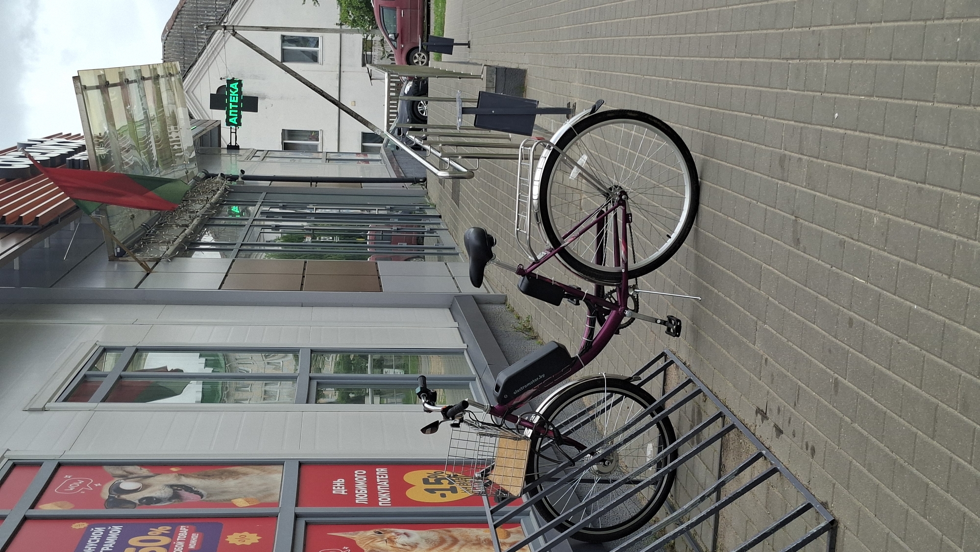 A purple electric bicycle is parked in a bicycle rack outside a store in Smalyavichy, Belarus. The bicycle has a black battery pack mounted on its frame and a basket on the front. In the background, a store with large windows displays advertisements, including one with a dog wearing sunglasses and another with a cat. A green sign with the word "АПТЕКА" (Pharmacy) is visible on the side of the building. The pavement is made of grey interlocking bricks, and there are metal railings and trash cans near the entrance of the store. The weather appears overcast.