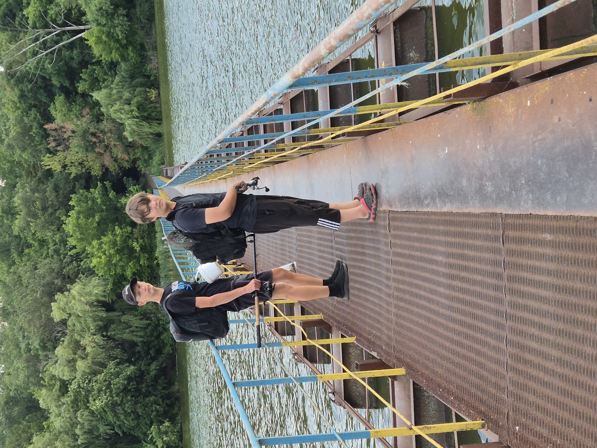 Two young males stand on a rusted metal walkway above water, which appears to be the dam of the Sozonivka reservoir in Sozonivka, Ukraine. The male on the left, wearing a black t-shirt, black shorts, black socks, and a black baseball cap, carries a black backpack and a white bucket. He is holding a fishing rod in his right hand. The male on the right, wearing a black t-shirt and black shorts with white stripes, carries a black backpack and is also holding a fishing rod. They are both facing the camera. The walkway is bordered by a blue and yellow railing. The background features a dense line of green trees and vegetation along the water's edge. The water is choppy and a greenish-blue color.