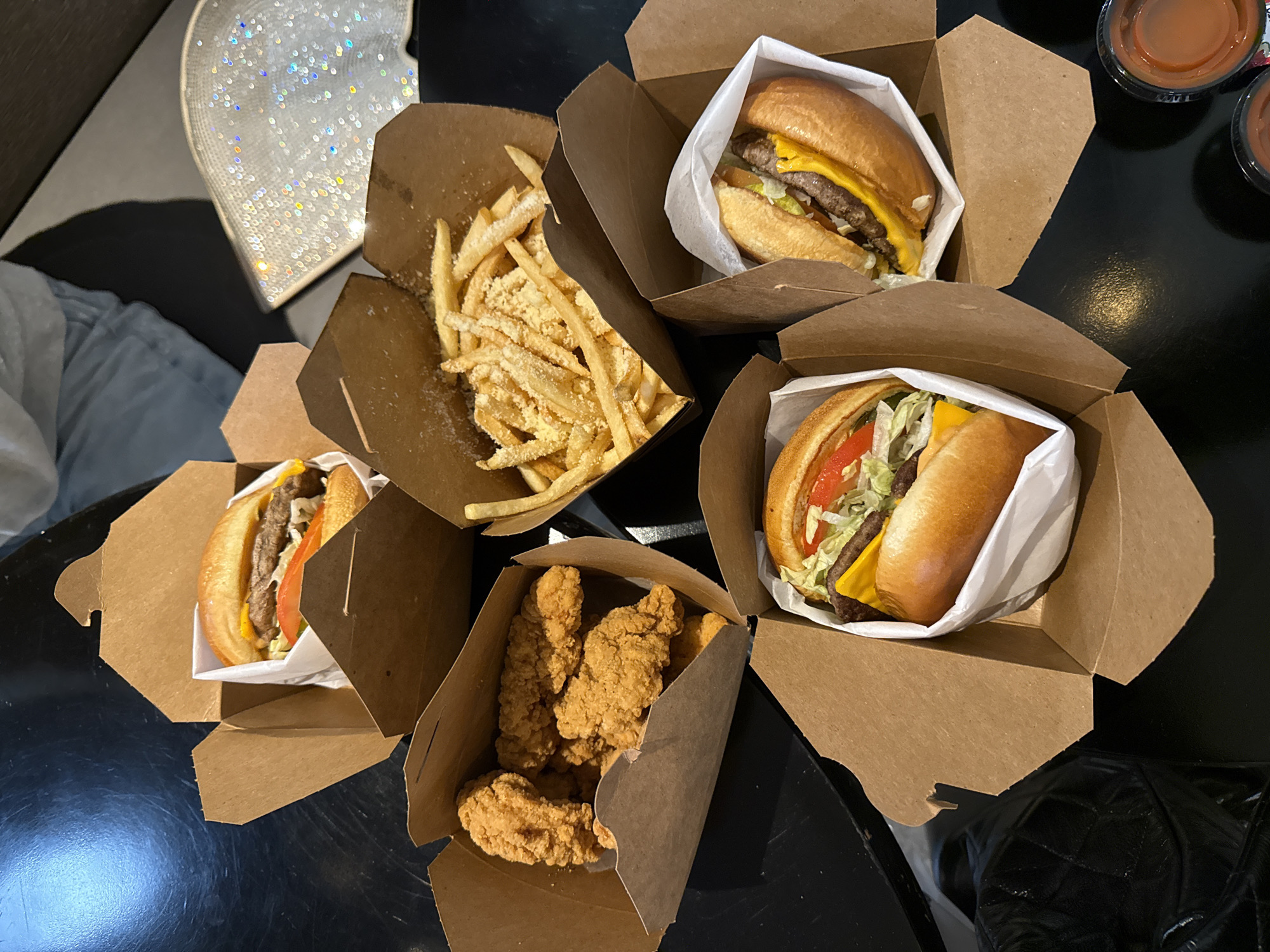 A high-angle shot depicts a collection of takeout food containers arranged on a dark, likely black, surface. The containers are cardboard, seemingly typical of fast-food packaging.
Three of the containers hold hamburgers, each with visible layers of bun, cheese, meat patty, lettuce, and tomato. Another container holds a serving of french fries, appearing seasoned with what looks like parmesan cheese. The fifth container holds fried chicken strips. Two small condiment containers are also visible.
The setting appears to be indoors, possibly a table at a restaurant or in a home. No individuals are visible in their entirety, but partial views of clothing (jeans, a white garment) suggest people are present. A sequin-covered object is also in view to the upper left.
Given the presence of In-N-Out style burgers with cheese, it is very likely that this scene takes place in Los Angeles. The brand is native to the area.