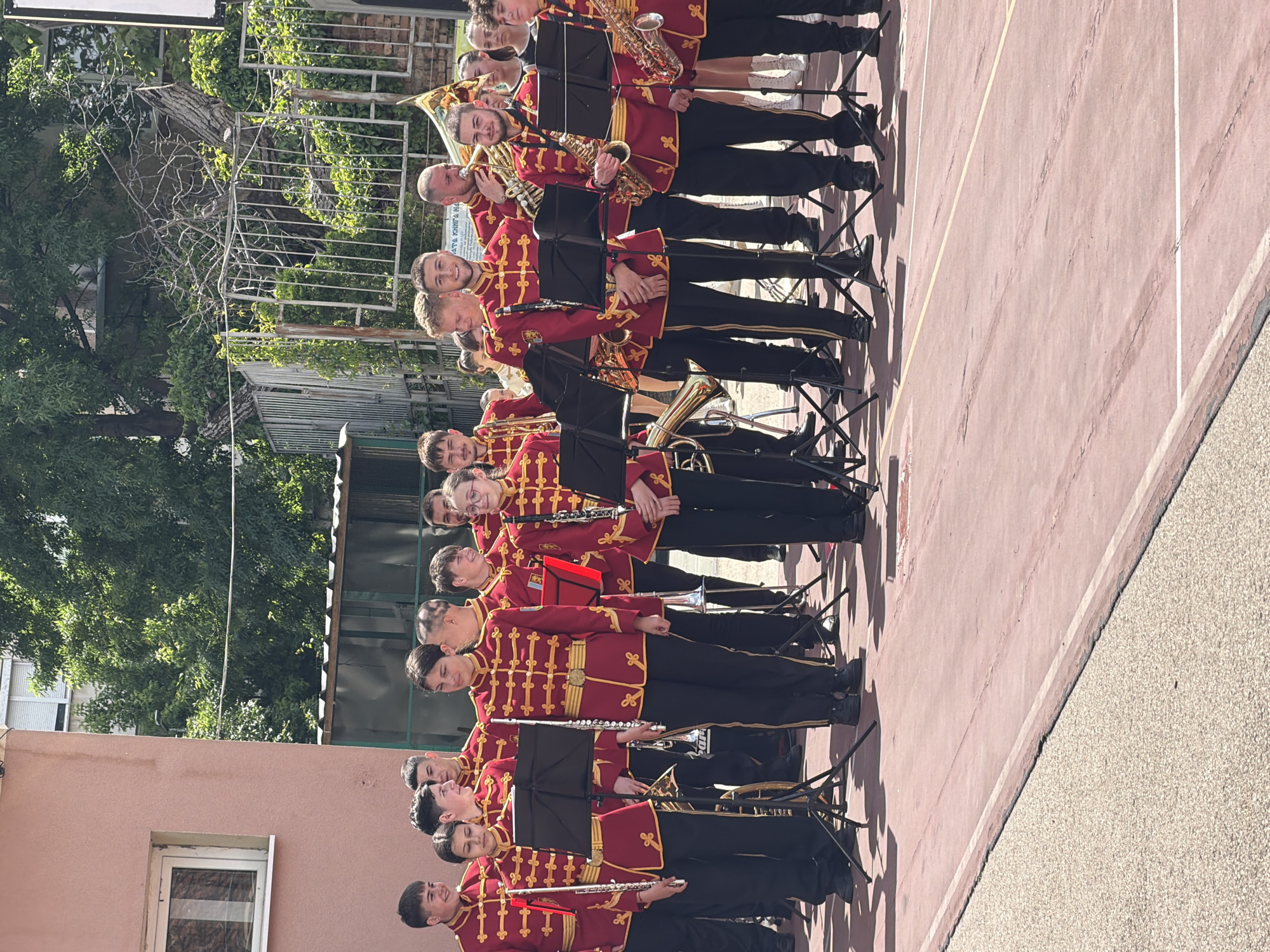 A group of people wearing red and gold military-style uniforms are arranged in rows on a red and white-lined surface, likely a track or a sports court. They are holding various musical instruments, including flutes, clarinets, trumpets, and saxophones. Some have black music stands in front of them. They seem to be standing at attention and possibly waiting to perform.
In the background, there is a building with a window and a fence. Trees with green leaves are visible behind the fence, indicating the presence of vegetation. The scene appears to be outdoors, with sunlight present. The location in the city of Pleven, Bulgaria is not evident from the image.