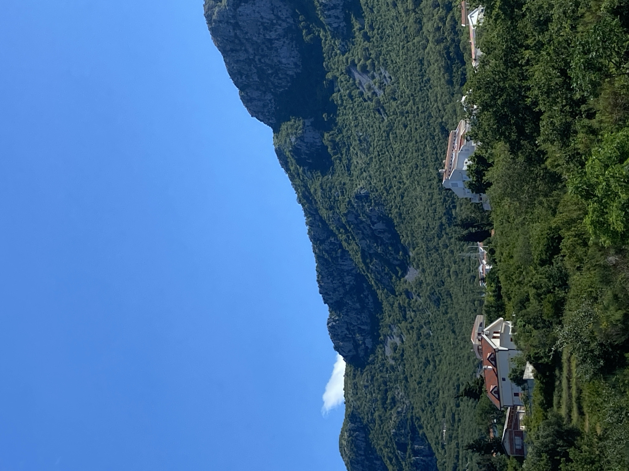 The image depicts a mountainous landscape with a village nestled at the foot of the mountains. The sky is a clear, vibrant blue with a single wisp of white cloud visible on the left. The mountains are covered in lush green vegetation, with rocky outcrops and steeper slopes visible. In the foreground and mid-ground, several houses with red tiled roofs are scattered amongst the greenery. The houses appear to be of modern construction with white or light-colored walls. Some trees and bushes are also present in the foreground, creating a sense of depth. The overall scene is one of a peaceful rural setting within a mountainous region. The location is identified as Budva, Montenegro.