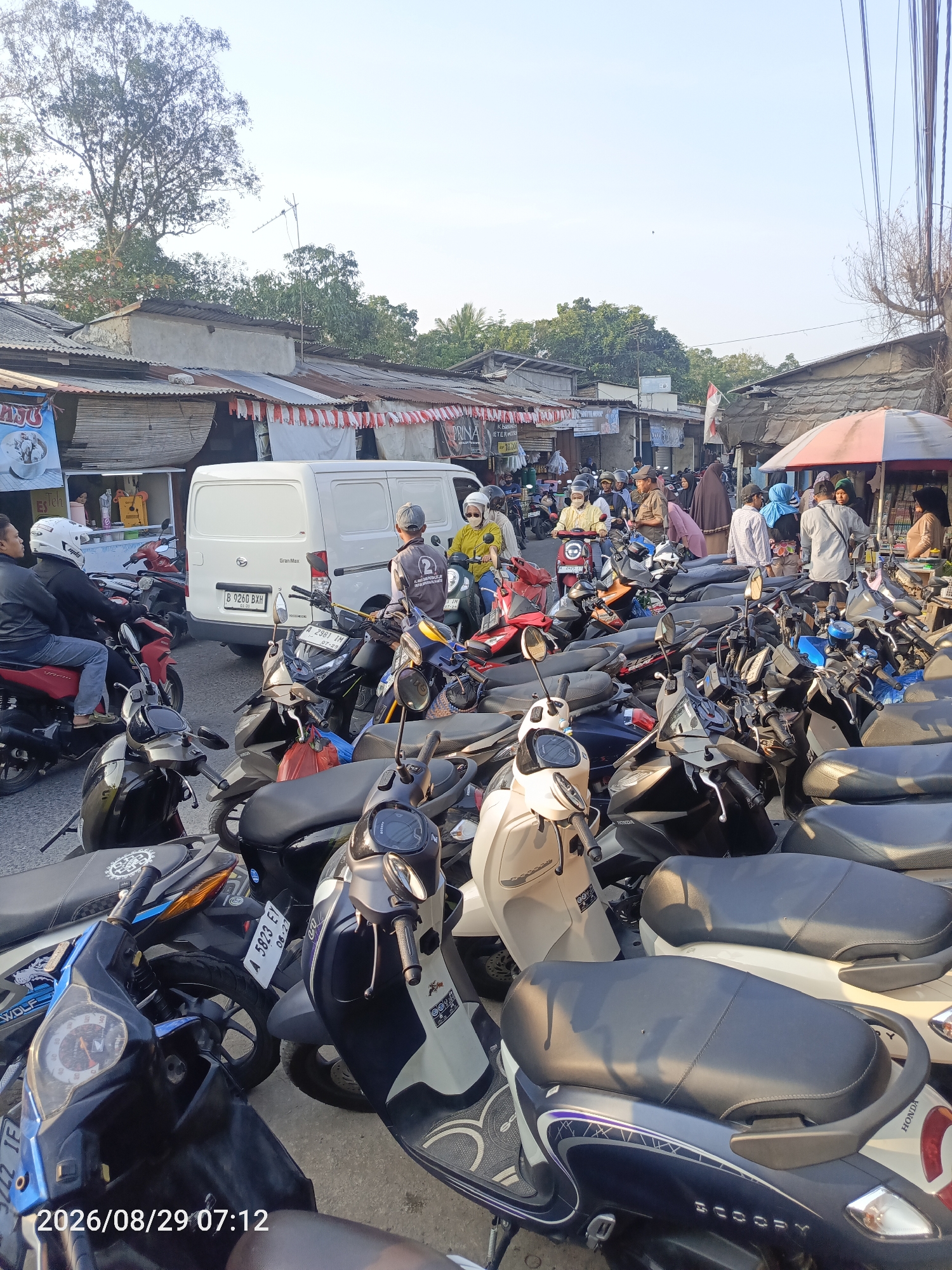 The image depicts a busy street scene in Sangiang, Indonesia, characterized by a dense arrangement of parked motorcycles. In the foreground, several scooters are visible, some with their seats exposed. Behind them, a white van with the license plate "B 9260 BXH" is parked, indicating a common mode of transportation. A variety of people, mostly dressed in casual attire, are interacting with the motorcycles, some appearing to be owners preparing to depart or having just arrived. The background reveals a bustling marketplace with small shops and stalls, many with corrugated metal roofs, typical of urban Indonesian street markets. The presence of red and white striped decorations, possibly flags or bunting, suggests a festive or nationalistic element to the scene. The overall atmosphere suggests a lively and active commercial area with a high volume of motorcycle traffic. The date stamp at the bottom indicates the image was taken on August 29, 2026, at 07:12.