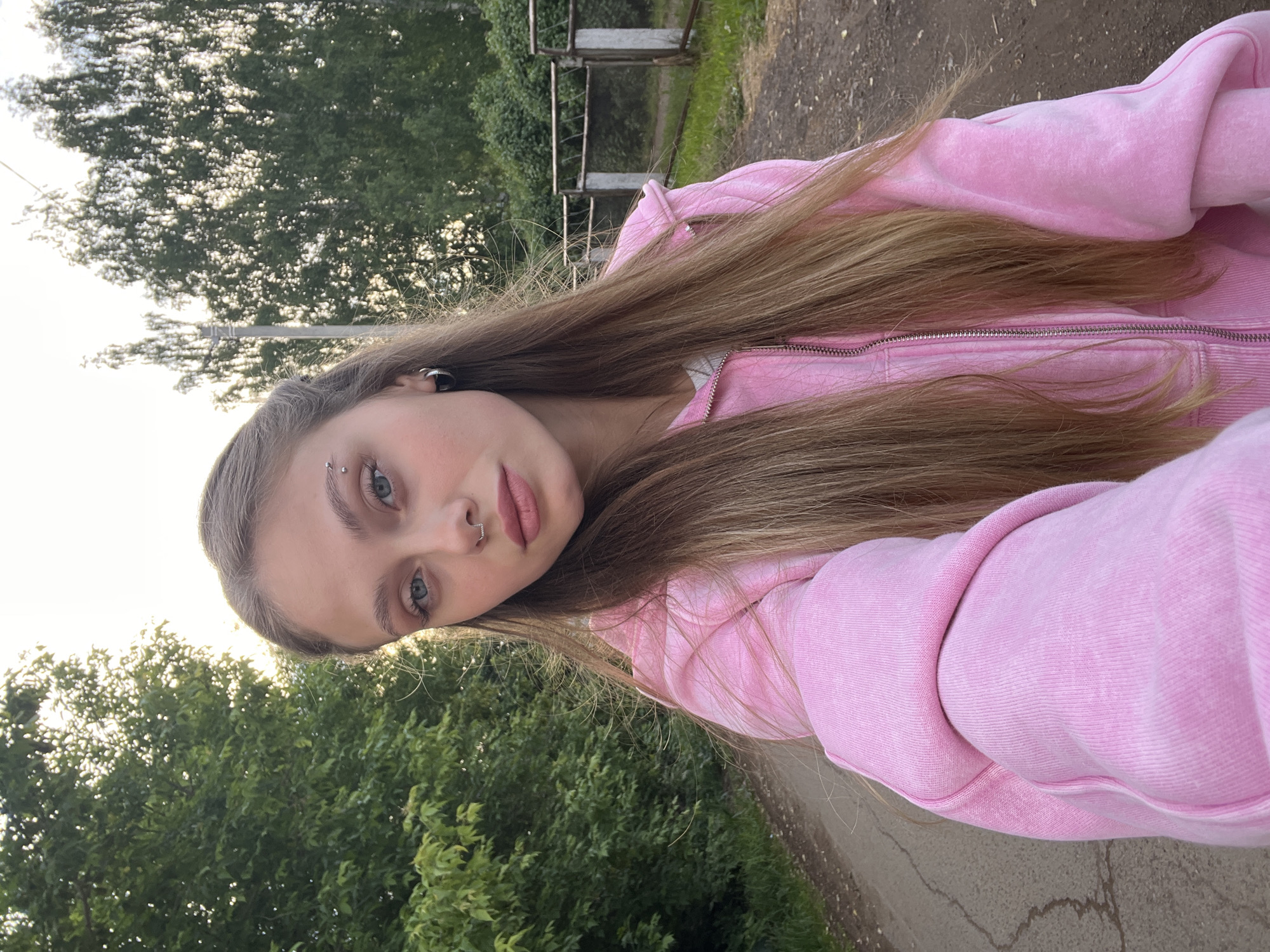 A young woman is posing for a selfie in an outdoor environment. She has long, light brown hair, light eyes, and pierced eyebrow, nose, and ear. She is wearing a pink zippered sweatshirt. The background consists of trees and a paved path or road. A metal fence is partially visible behind the trees. The location, Фоки, Russia, cannot be determined with certainty from the image content.