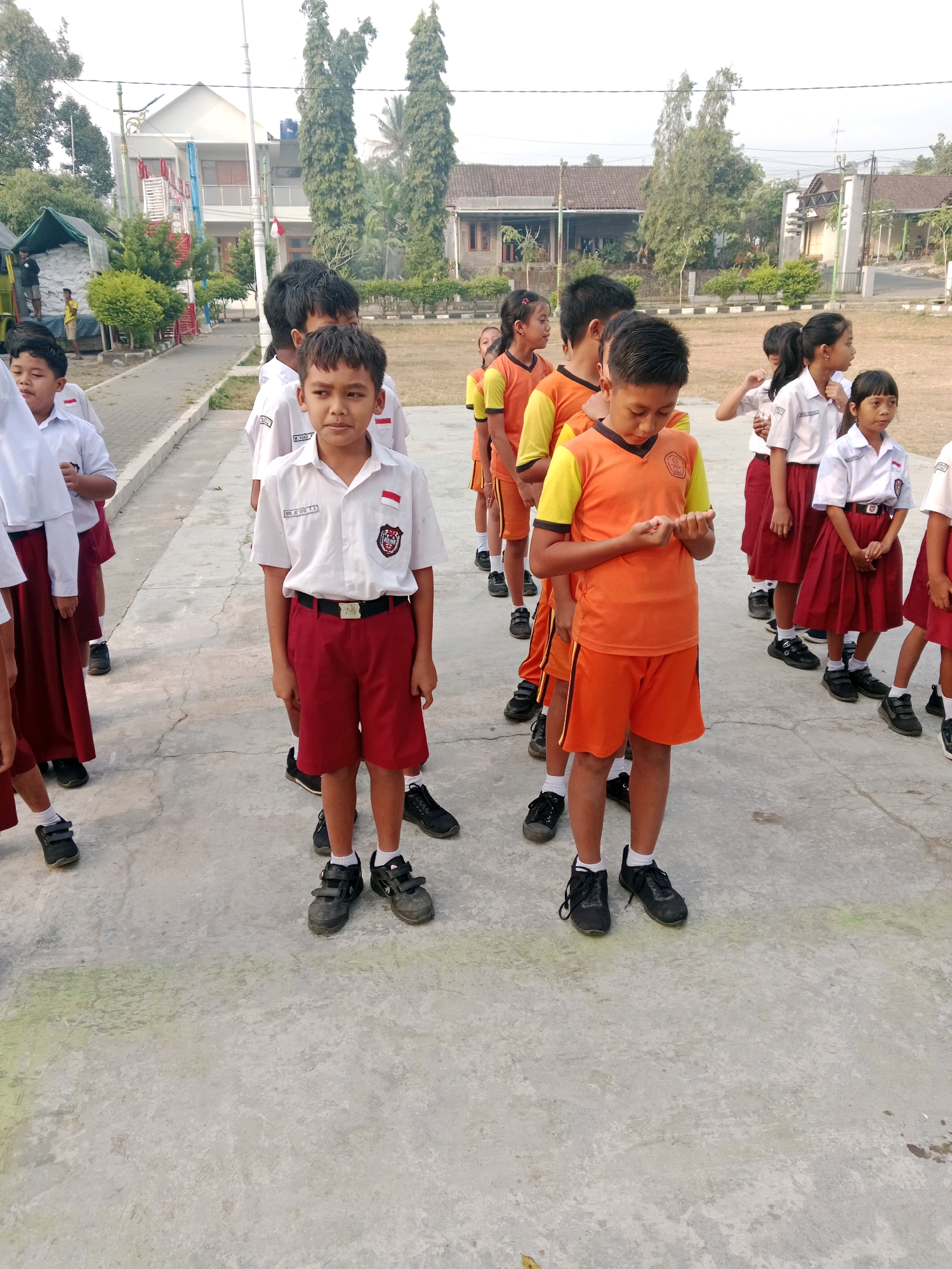 In Salatiga, Indonesia, a group of elementary school students stand in a line outdoors. The children are wearing their school uniforms: white shirts with red trim and patches, and either red shorts or red skirts. The boy in the foreground, looking directly at the camera, wears a white short-sleeved shirt with a crest on the left breast and red shorts. He has black shoes and socks. Behind him, other students are visible in a line, some in similar uniforms, others in orange and yellow shirts with black shorts. The background shows a building with white walls and a tiled roof, surrounded by trees and greenery. The ground is paved with concrete.