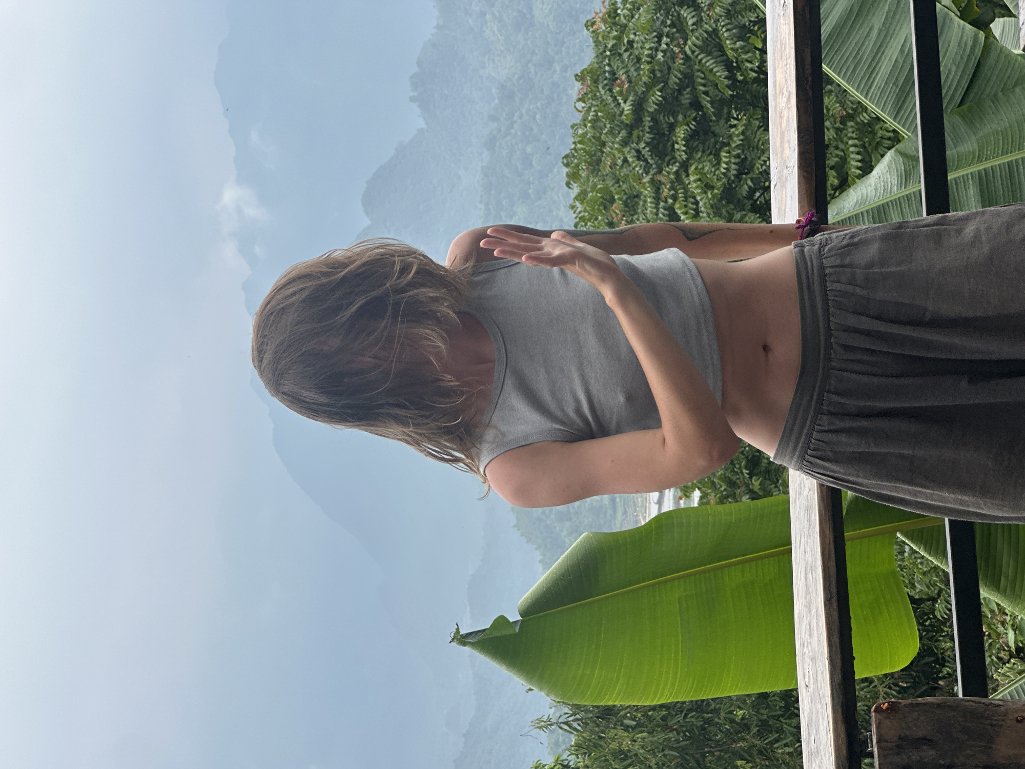 A woman stands on a wooden balcony overlooking a lush, mountainous landscape in Pù Luông Commune, Vietnam. Her face is obscured by her long, light brown hair. She is wearing a grey crop top and loose grey pants. Her right arm is extended forward, with her palm facing outwards as if to stop something or gesture. To her left, a large, vibrant green banana leaf is prominent. The background features rolling hills covered in dense vegetation and mist, suggesting a humid, possibly rainy, day. A wooden railing runs across the bottom of the frame, separating the balcony from the scenic view.