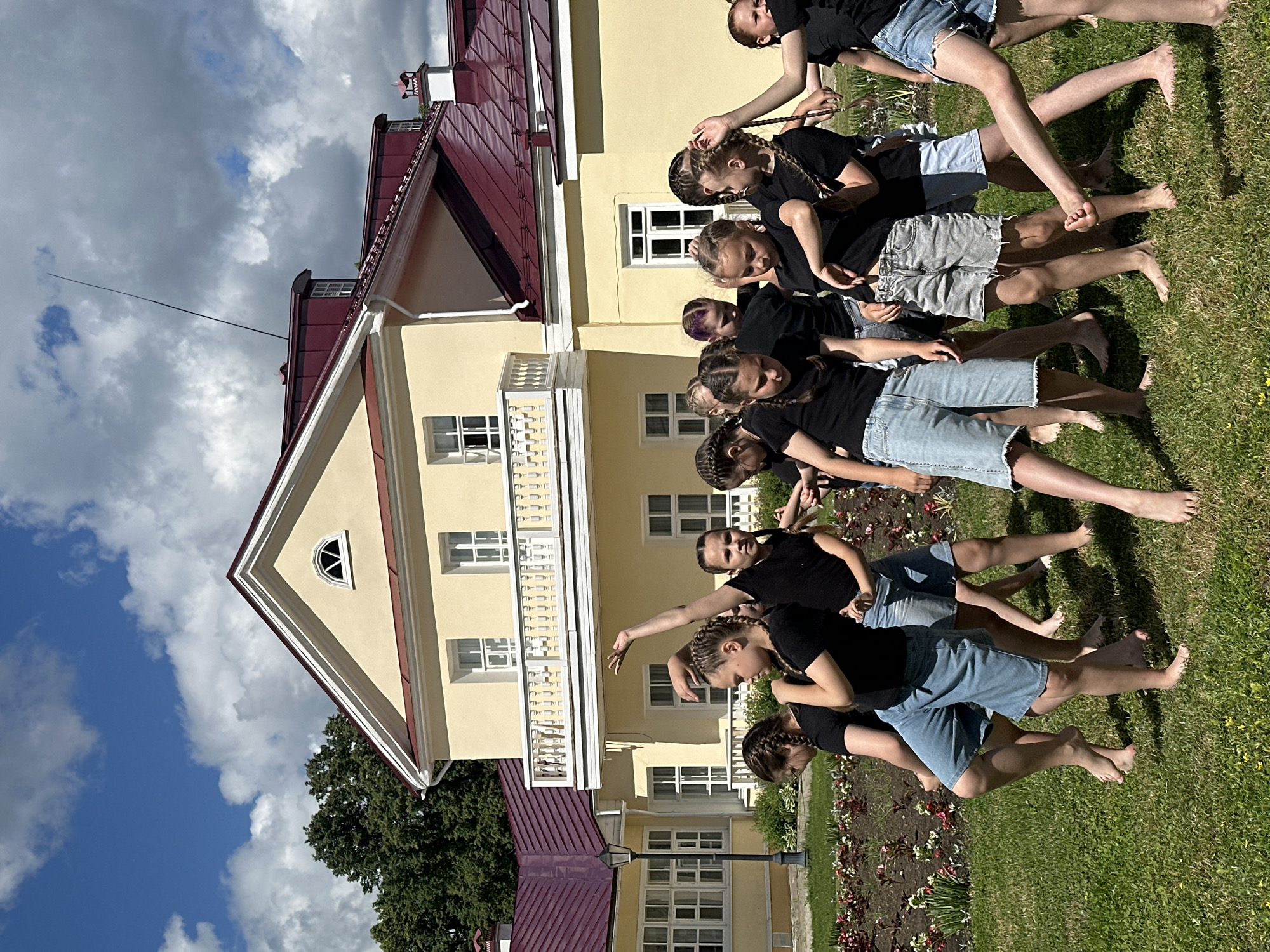 A group of young girls, dressed in black tops and denim shorts, are performing an outdoor dance rehearsal in the city of Votkinsk, Russia. They are barefoot and appear to be mid-motion, with some arms raised and bodies in dynamic poses. The background features a yellow building with a red roof, characteristic of a historic estate, possibly the Votkinsk State Historical-Architectural Museum-Reserve, which was the birthplace of composer Pyotr Ilyich Tchaikovsky. A manicured garden with flower beds and green grass surrounds the performers, under a partially cloudy sky. The scene conveys a sense of youthful energy and artistic activity within a cultural landmark.