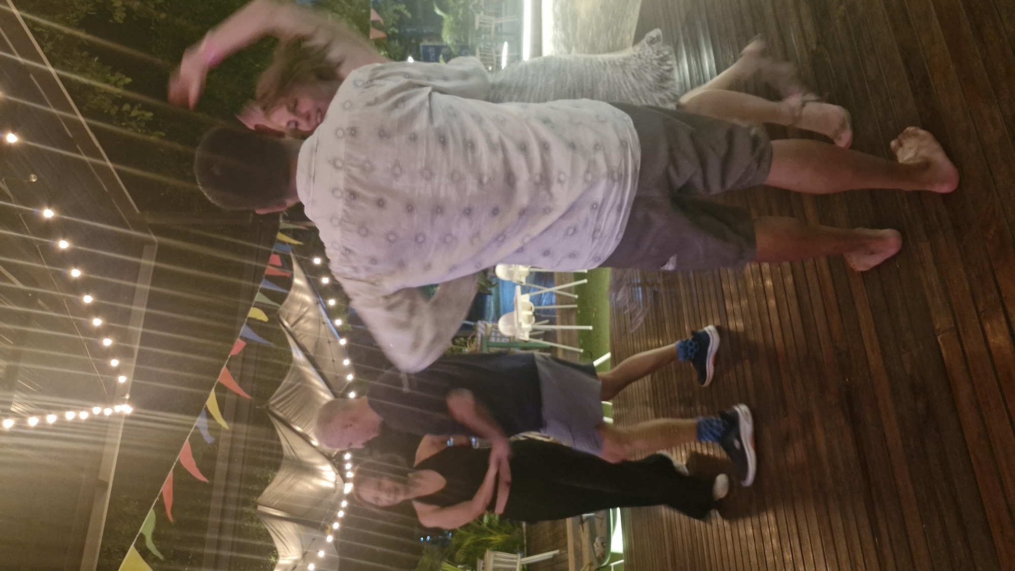 The image shows four people dancing at what appears to be an outdoor event at night. The scene is lit by strings of warm, glowing lights and colorful triangular flags that are strung across the space. The ground is made of wooden planks, suggesting an outdoor patio or deck.
In the foreground, two people are dancing. One person, wearing a black tank top and black pants, is being held by another person wearing a dark t-shirt and grey shorts. The couple in the foreground are barefoot.
Behind them, two other people are also dancing. One person, wearing a light-colored patterned shirt and grey shorts, is holding another person, who is wearing a light-colored, possibly white, dress. This couple appears to be barefoot as well.
The overall mood of the image is energetic and festive, suggesting a party or a social gathering. The time of day is clearly night due to the artificial lighting. There is no visible text in the image.