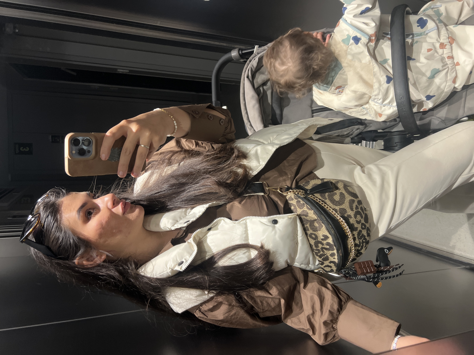 An adult female and a young child are depicted in an indoor setting, likely a mirrored elevator or hallway, within Kiryat Bialik, Israel. The female, with long dark hair and sunglasses perched on her head, is taking a mirror selfie with a brown-cased iPhone held in her right hand. She wears a brown long-sleeved shirt, a cream puffer vest, and light-colored pants. A leopard-print fanny pack, adorned with keychains resembling a chocolate bar and a black duck, is slung across her body. A child with light brown, curly hair is seated in a grey stroller to the female's right, facing away from the camera. The child wears a cream-colored hooded garment patterned with abstract blue, orange, and green shapes. The background consists of dark, reflective surfaces.