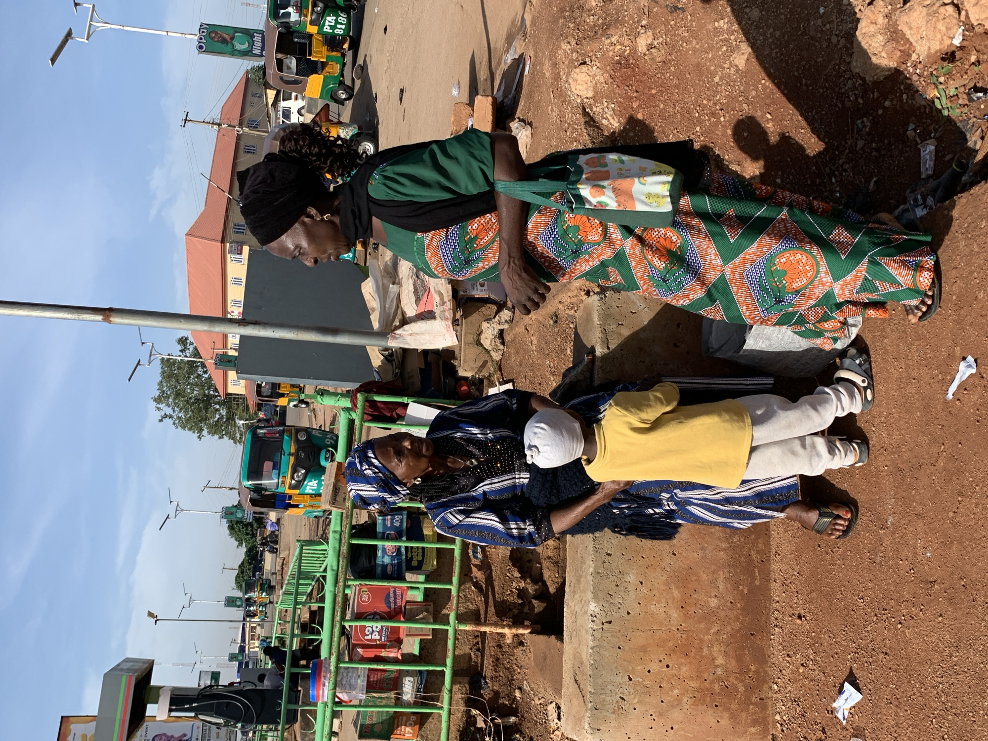The image captures a sunlit street scene, likely in Gura Top, Nigeria, featuring two women and a child. The ground is reddish-brown and dusty, characteristic of the region.
On the right side of the frame, a woman with dark skin is seated on the ground. She wears a black headscarf, a green long-sleeved top, and a vibrant patterned skirt in shades of green, orange, blue, and white. A white bag adorned with colorful cartoon animals rests on her lap. Her bare feet are visible, wearing dark sandals.
To the left, another woman sits, dressed in a blue and white striped or patterned outfit with a dark head covering. Beside her, a child sits or leans, wearing a yellow long-sleeved top, light grey pants, and a white cap. The child also wears sandals.
In the background, a busy street scene unfolds. A green metal barrier or fence runs along the roadside, behind which various packaged goods are visible, including boxes marked "POP" and a product resembling "INDOMIE" noodles. Several green and yellow tricycles, commonly known as keke napep, are parked or navigating the street. One tricycle clearly displays "PTA-0186" on its back. Overhead, modern street lights are mounted on poles. A banner advertisement for "Opay" is visible on one pole, featuring a person's image and the word "Night." Beyond the street, buildings with reddish-brown roofs can be seen under a clear, bright blue sky. Strong shadows indicate bright daylight, suggesting mid-day or early afternoon. The women and child appear to be resting or waiting by the roadside in this bustling environment.