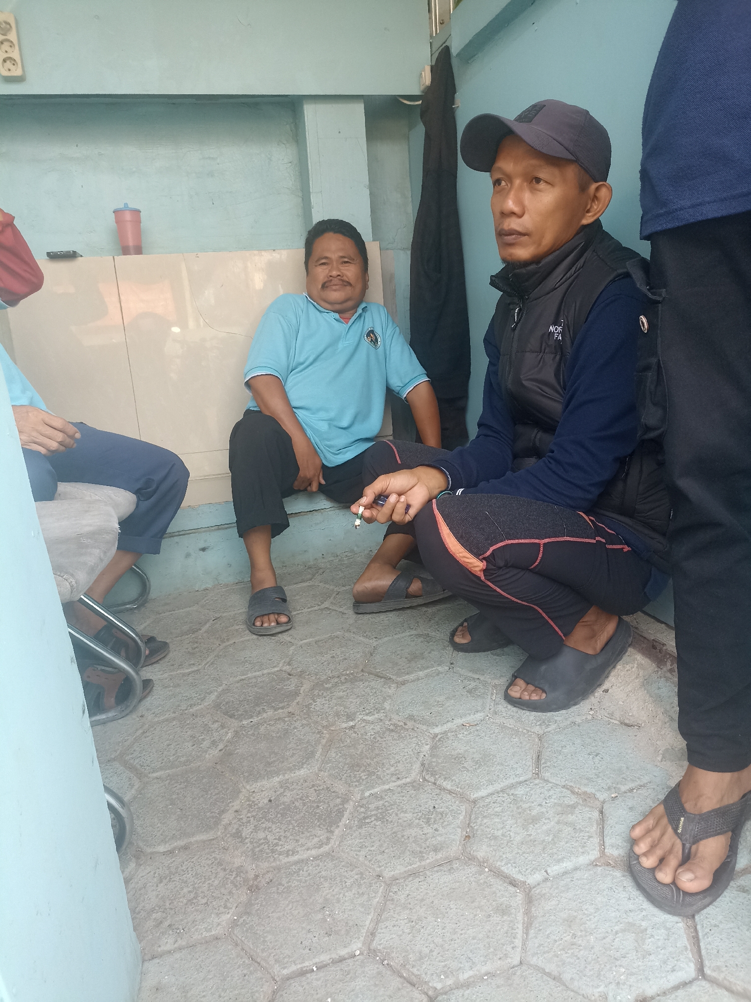 In Cibarusah, Indonesia, three men are seen gathered outdoors. One man is seated on a low wall, wearing a light blue polo shirt and dark pants. Another man, wearing a dark vest over a blue long-sleeved shirt and a dark baseball cap, is crouched down, holding a cigarette. The third man, partially visible, is seated on a chair or bench, wearing a blue shirt and dark pants. The ground is paved with hexagonal tiles. The background shows a light blue wall and a beige counter with a red cup and a black object on it.