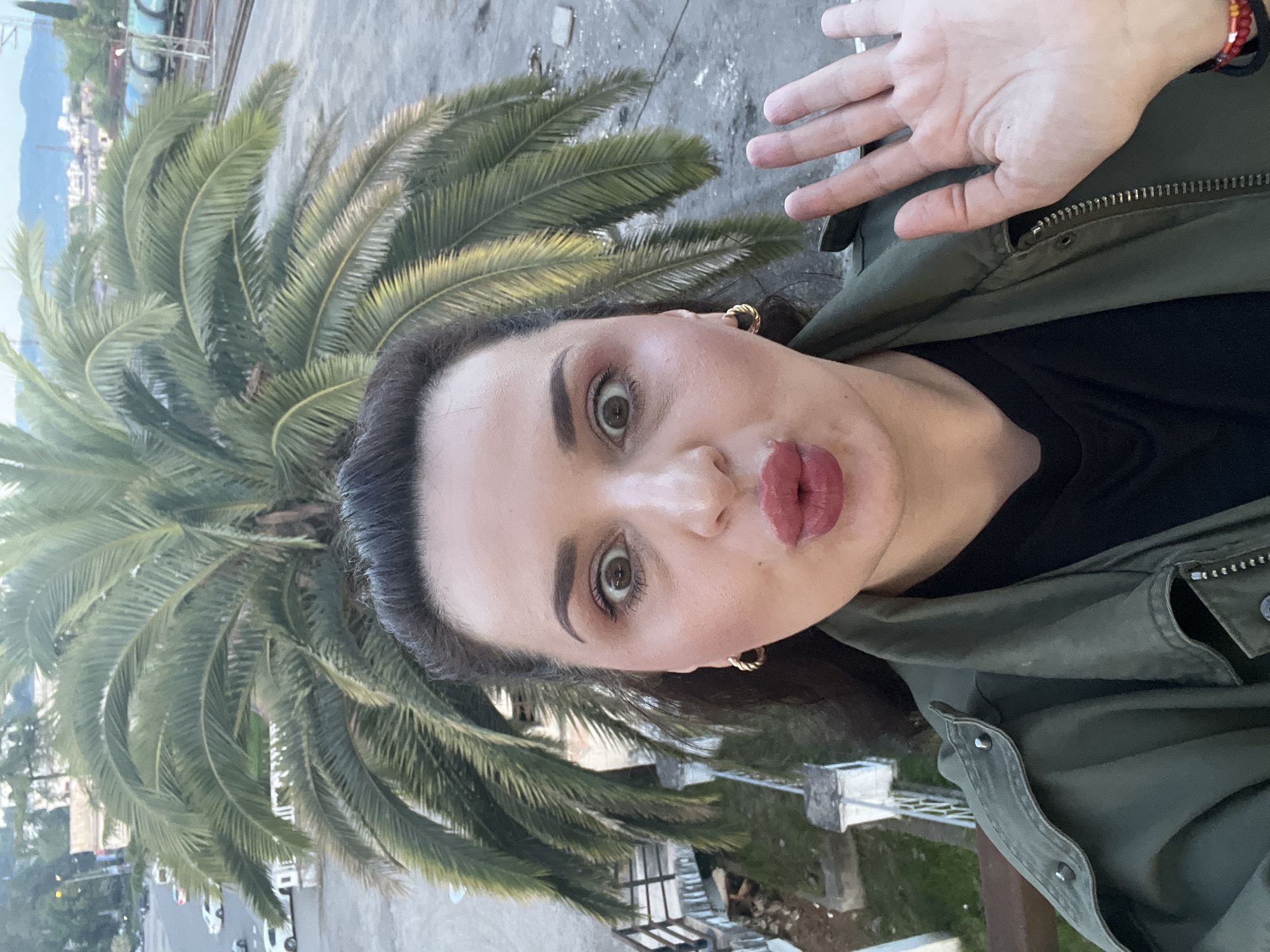 A medium close-up selfie shows a woman with dark hair and a light complexion, angled slightly to her left, with her gaze directed towards the camera. Her eyes are wide open, and her lips are pursed in a 'kiss' or 'duck face' expression. She has noticeable makeup, including eyeliner, eyeshadow, and lipstick. She wears a black top underneath an olive green jacket with a visible zipper and buttons. A gold hoop earring is visible on her right ear. Her right hand is raised, palm open, extending into the upper right corner of the frame. On her right wrist, red and black bracelets are visible.
The background features a large palm tree with lush green fronds, prominent on the left side of the image. Beyond the palm tree and to the left, distant mountains or hills are visible under a bright sky, suggesting a coastal or lakeside setting. Below and to the right of the palm tree, urban elements like buildings, a street with parked cars, and railings or balcony structures can be seen, indicating an elevated vantage point, possibly from a balcony or window. The lighting suggests it is daytime, possibly late afternoon, with soft, ambient light. There is no visible text in the image.
