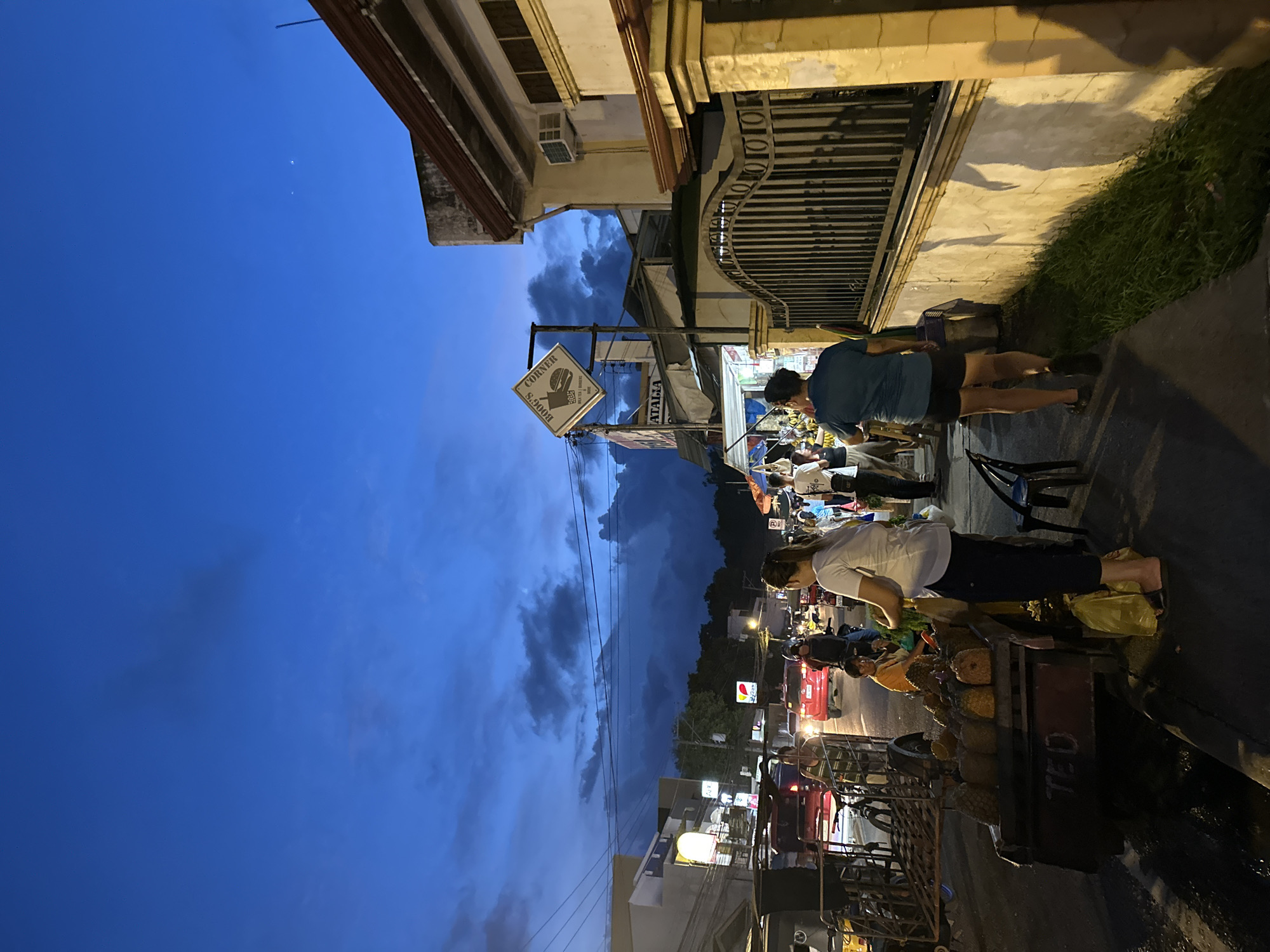 The image depicts a street scene in Davao City, Philippines, during dusk. The sky is a deep blue with dramatic clouds, suggesting the end of the day. Several people are walking along the sidewalk, some browsing at roadside stalls. A fruit vendor's cart is prominently displayed in the foreground, laden with pineapples. Vehicles, including cars and motorcycles, are visible on the road, indicating traffic. Buildings line the street, some with illuminated signs, suggesting commercial activity. A sign for "BOOG'S CORNER" is visible, along with other signage that is partially obscured. The overall atmosphere is that of a bustling local market or commercial area at twilight.