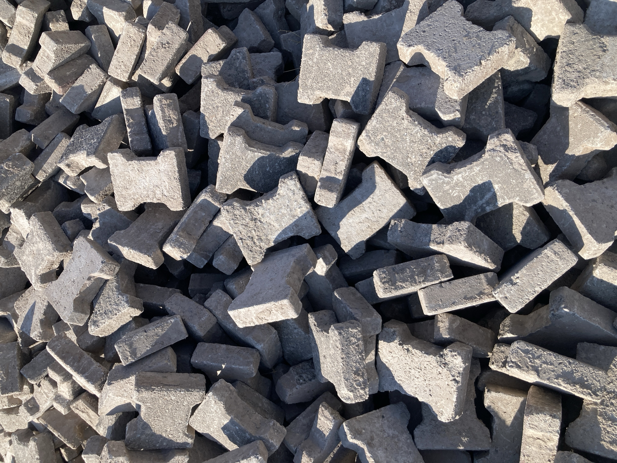 A pile of interlocking concrete pavers, also known as "I" pavers, are stacked outdoors in an area that appears to be a construction or landscaping site in Avanos, Turkey. The pavers are a light gray color and show some wear and tear. The sunlight casts shadows across the pile, highlighting the texture of the concrete. There are no people or other objects visible in the image.
