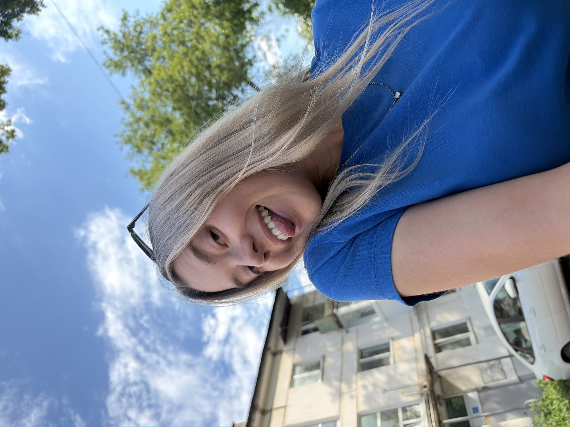 A young woman with blonde hair and a bright blue t-shirt takes a selfie outdoors. She is sticking her tongue out playfully at the camera, her mouth open in a wide smile. She wears a delicate necklace with a small pendant. In the background, a light-colored apartment building with multiple windows and balconies stands under a blue sky with scattered clouds. A white sedan is parked in front of the building. Green trees are visible to the right of the frame. The perspective is from a low angle, looking up at the woman and the sky. The scene appears to be in a residential area of the city of Chaikovsky, Russia.