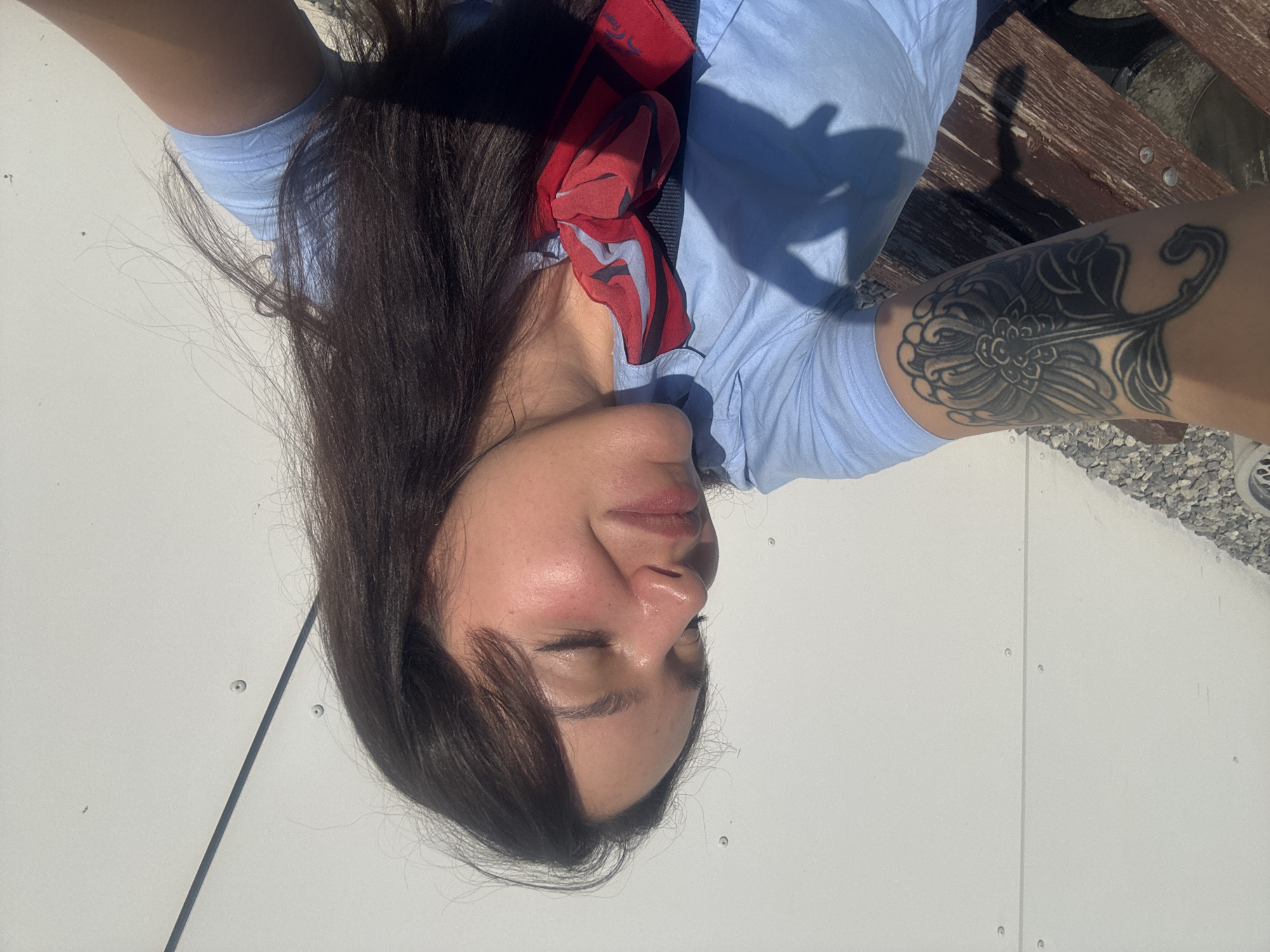 A young woman is captured in a selfie outdoors. She has long, dark brown hair and her eyes are closed. She is wearing a light blue collared shirt with a red scarf tied around her neck. A large, black floral tattoo is visible on her left forearm. The background consists of a white corrugated metal wall and a wooden bench with gravel in the foreground. The lighting suggests it is daytime, with sunlight casting shadows. The scene appears to be in Burgas, Bulgaria, possibly near an industrial or coastal area given the wall and gravel.