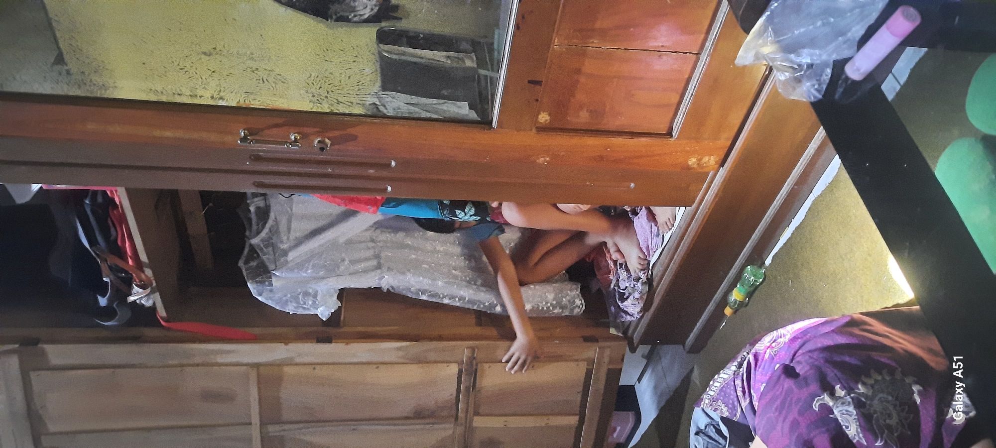 An undated, low-angle, full-shot, indoor, eye-level photograph taken in Ploso, Indonesia, depicts a young child crouched inside an open wooden wardrobe. The child, whose face is obscured by their body, is wearing a blue top and patterned shorts, and is surrounded by clothes and fabric. Their left arm is extended towards the side of the wardrobe, and their legs are bent. The wardrobe, made of dark wood, contains various garments, including a white dress on a hanger. Outside the wardrobe, a purple and white patterned fabric is visible on the floor in the foreground, and a green bottle is on the floor. The background wall is textured yellow. The lighting is dim, with a bright light source illuminating the floor area near the bottle. The photograph is attributed to a Galaxy A51.