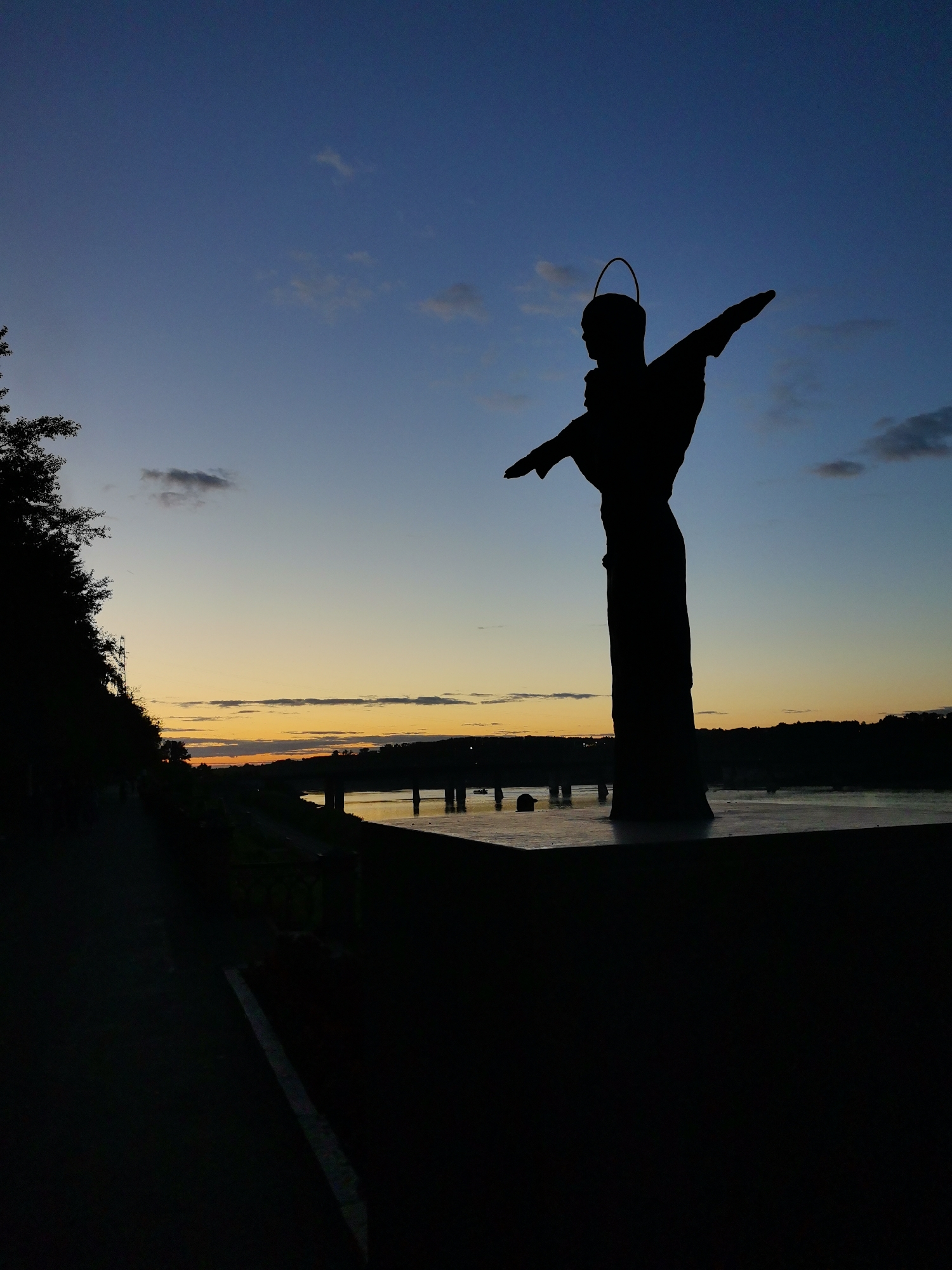 The image depicts a silhouette of the monument "To the Motherland" in Kemerovo, Russia, during twilight. The statue, resembling Jesus Christ with arms outstretched and a halo, stands prominently against a gradient sky transitioning from deep blue to orange hues near the horizon. In the foreground, a dark pathway or embankment runs along the left side, leading towards the river. The river itself reflects the warm colors of the sunset, and a bridge with illuminated supports is visible in the distance. The overall atmosphere is serene, with the silhouette emphasizing the monument's form against the natural beauty of the sunset.