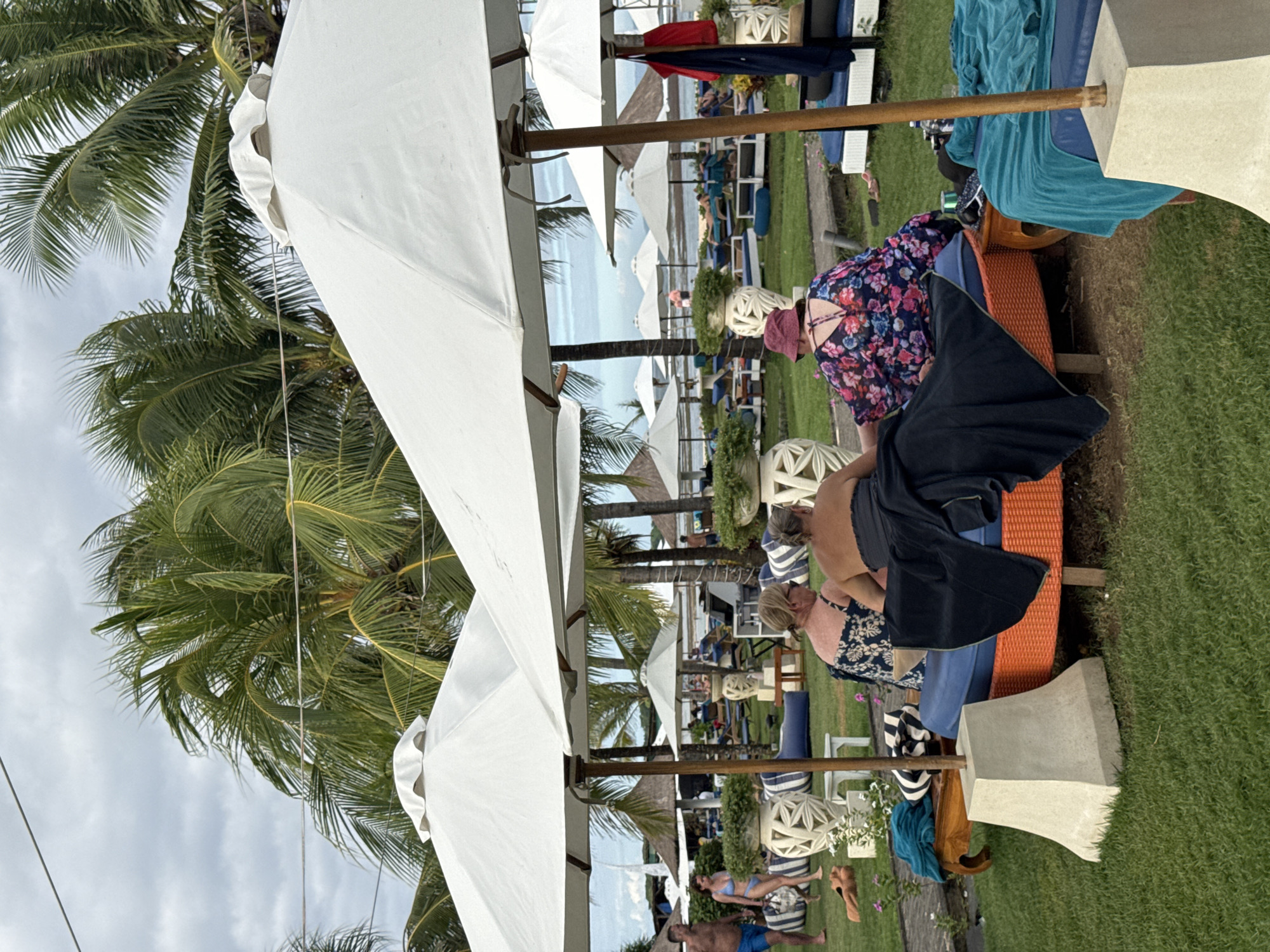 The image depicts a coastal resort scene, potentially in Kuta Selatan, Indonesia. White sun umbrellas dominate the foreground, suggesting a beach or pool area. Several people are seen relaxing on lounge chairs, some under the shade of the umbrellas. The sky is overcast. In the background, there is a glimpse of the ocean. Palm trees are visible adding to the tropical setting.