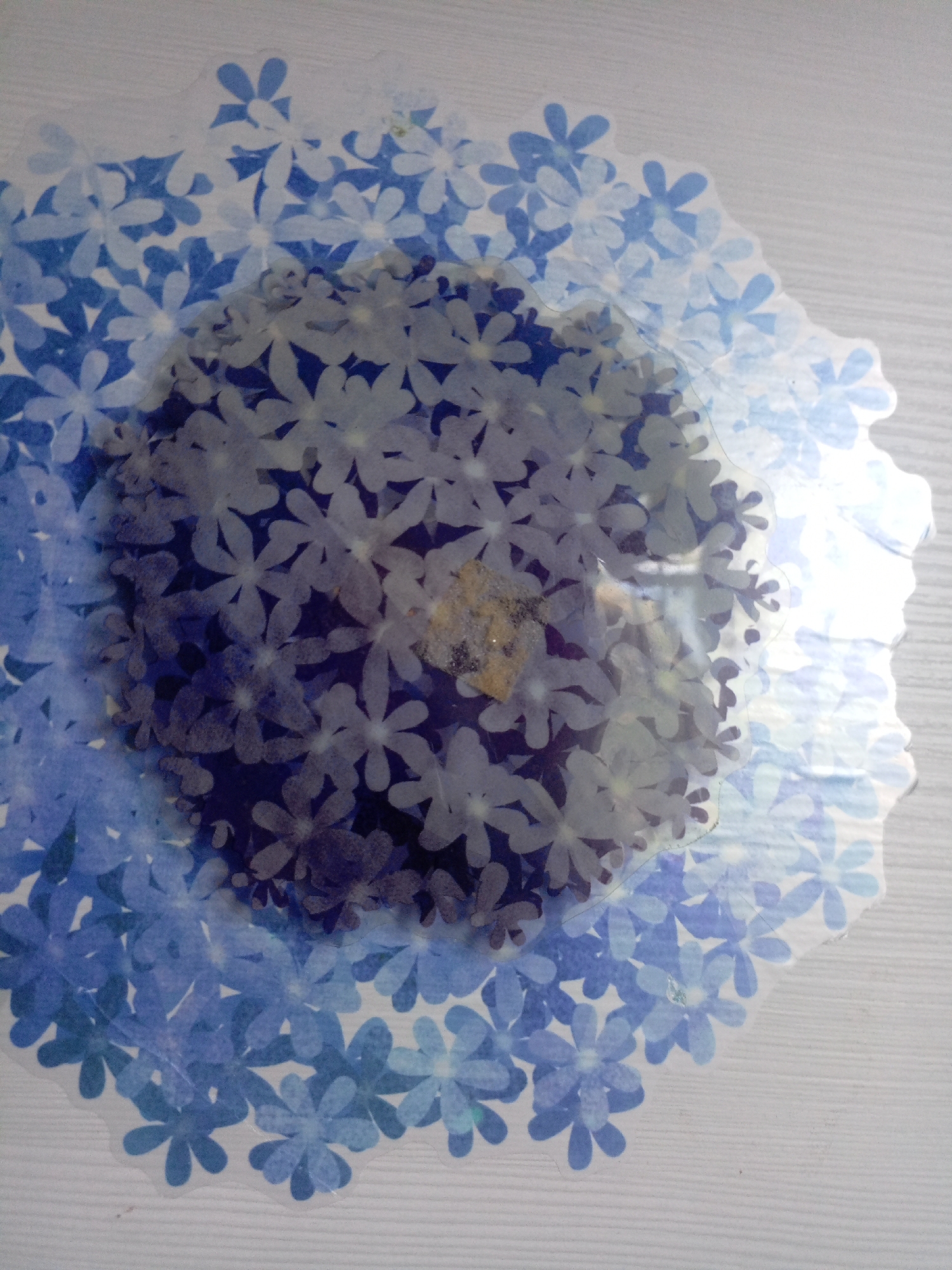 The image displays a close-up view of a circular design made of overlapping blue and white flower-shaped cutouts. The cutouts are layered, creating a sense of depth and a mandala-like pattern. The flowers vary in shade from deep indigo to pale blue and white. Some of the cutouts appear to be translucent, allowing the layers beneath to show through. The edges of the design are scalloped and irregular. The background is a plain, light-colored surface with a subtle wood grain texture, suggesting it might be a table or a wall. There are no people or animals visible in the image. The image does not contain information about the location in Veshki, Russia.