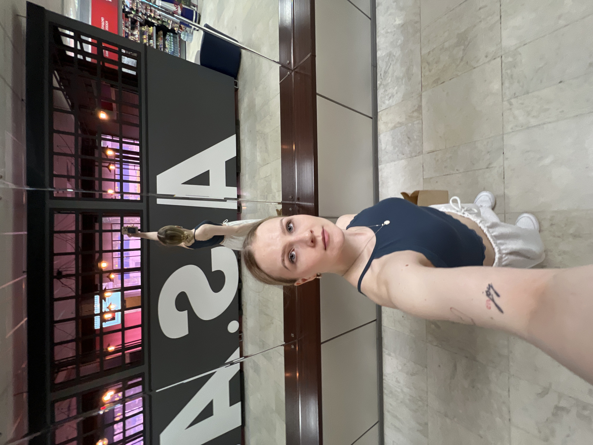 A selfie shows a white woman with blonde hair and fair skin. She is wearing a dark blue tank top and light-colored shorts with white sneakers. The woman is standing on a tiled floor and is positioned in front of a black wall with the number "42" in large white numerals. The background reflects a building with multiple floors, and within the building, decorative lighting is visible. The woman is holding the camera in her outstretched right arm. There is a tattoo on the right arm. The location is in Khimki, Russia.