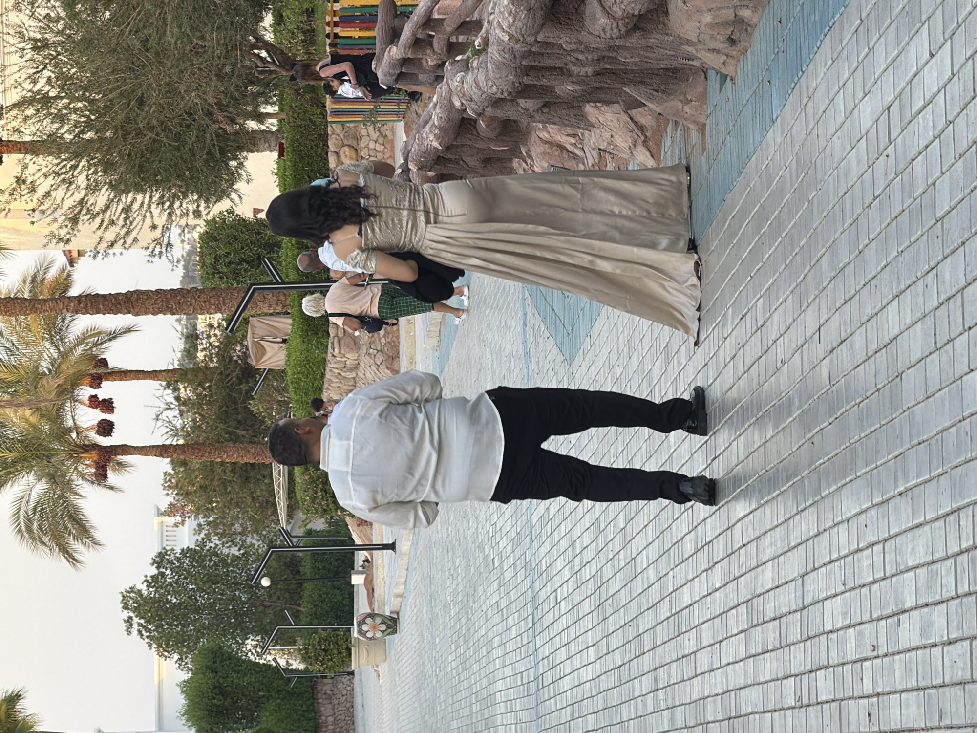 In Sharm El Sheikh, Egypt, an outdoor paved walkway features a man in a white long-sleeved shirt and black trousers walking away from the camera, his back to the viewer. Beside him and slightly ahead, a woman in a shimmering champagne-colored, off-the-shoulder evening gown also walks away from the camera. Behind them, a couple in casual attire, a woman with blonde hair in a pink top and green pants, and a man in a light blue shirt, are also walking. Further in the background, on a colorful fence, two women are visible, one in a black dress and the other a person who might be a child. Palm trees with clusters of dates are scattered throughout the scene, alongside well-maintained greenery and decorative lighting fixtures. A stone retaining wall with a rustic, wooden-like railing borders the walkway on the right. The ground is paved with rectangular gray tiles, interspersed with a strip of blue tiles. The overall environment suggests a resort or recreational area in Sharm El Sheikh.