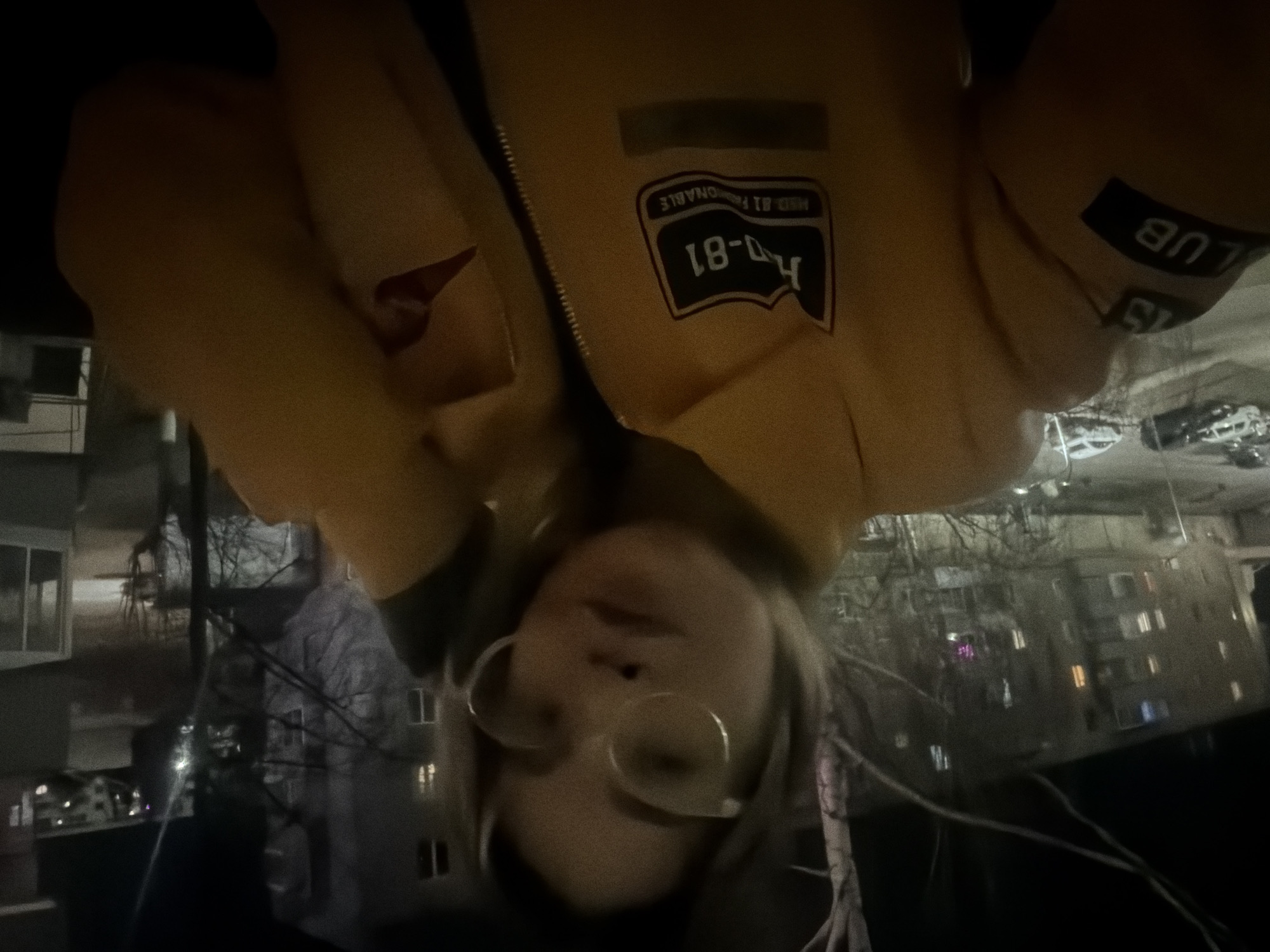 A selfie taken from a high angle shows a person wearing round glasses and a yellow jacket with "H-81" and "CLUB 15" patches. Outside, it is dark, and apartment buildings are visible in the background. The location is Naberezhnye Chelny, Russia.