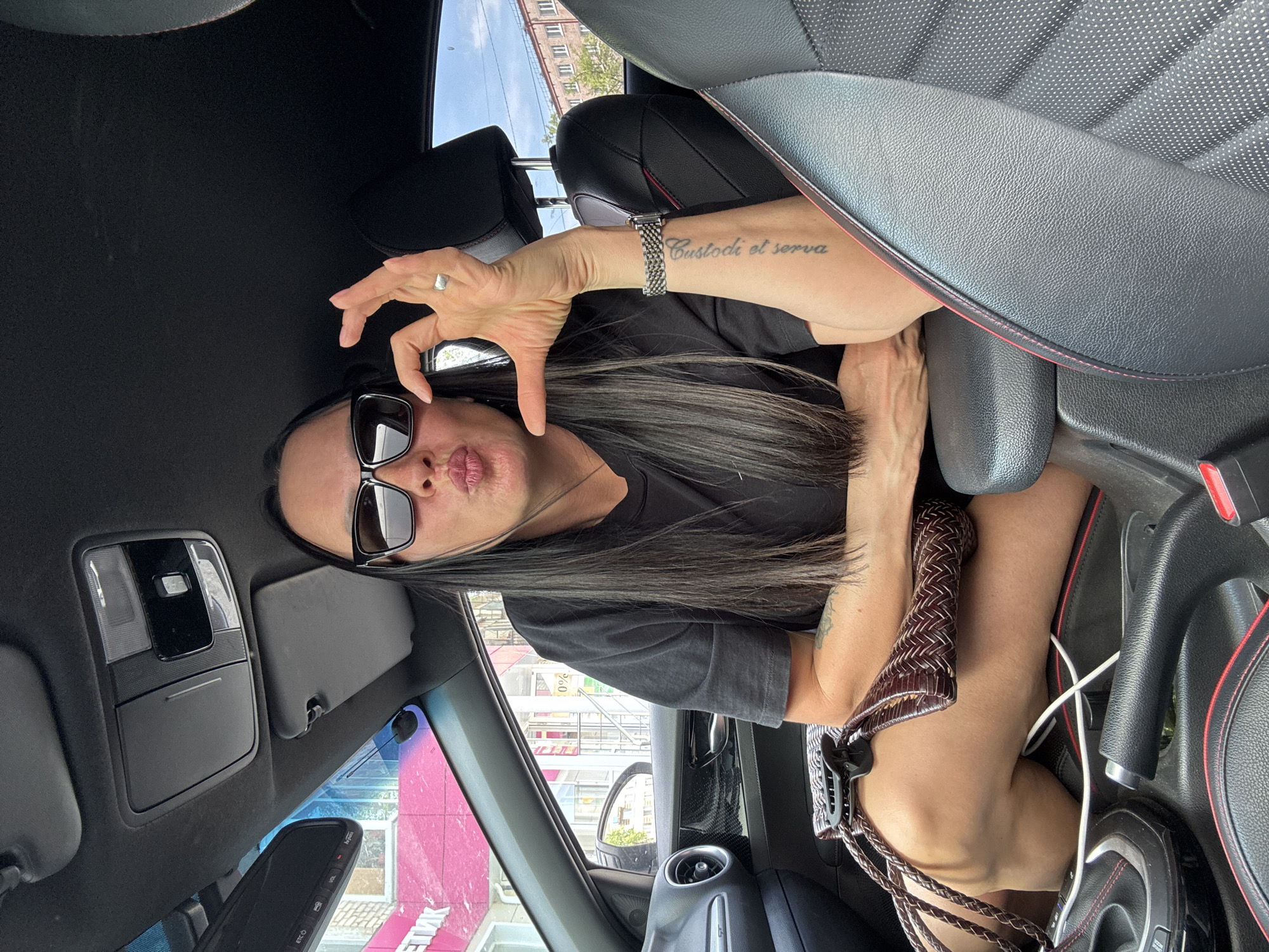 A woman is sitting in the driver's seat of a car, making a pouting face and holding up one hand. She is wearing sunglasses and a black t-shirt. The interior of the car is black with red trim. Through the car window, a pink building with Cyrillic text on it is visible. Based on the text on the building, the location is likely Chaikovsky, Russia. A brown woven purse sits in the center console.