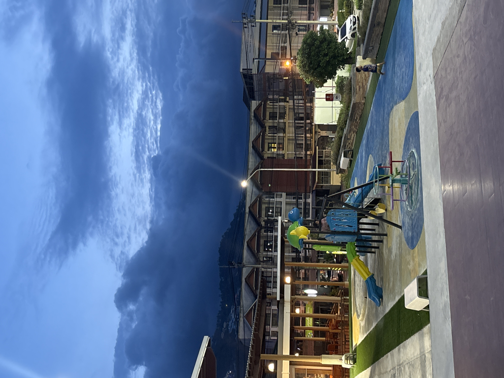 This photograph, taken at night, depicts a playground area in Baan Bo Phut, Thailand. The image is oriented vertically, with the sky occupying the left side of the frame. Dark, ominous clouds fill the sky.
The playground is constructed with a blue, yellow, and white color scheme. Playground equipment, including a slide and climbing structures, is visible. The ground is partially covered with artificial turf.
Adjacent to the playground, a multi-story building with lighted windows is visible. A street light illuminates the area.
A child is walking along a pathway within the playground area. A white vehicle is parked nearby.