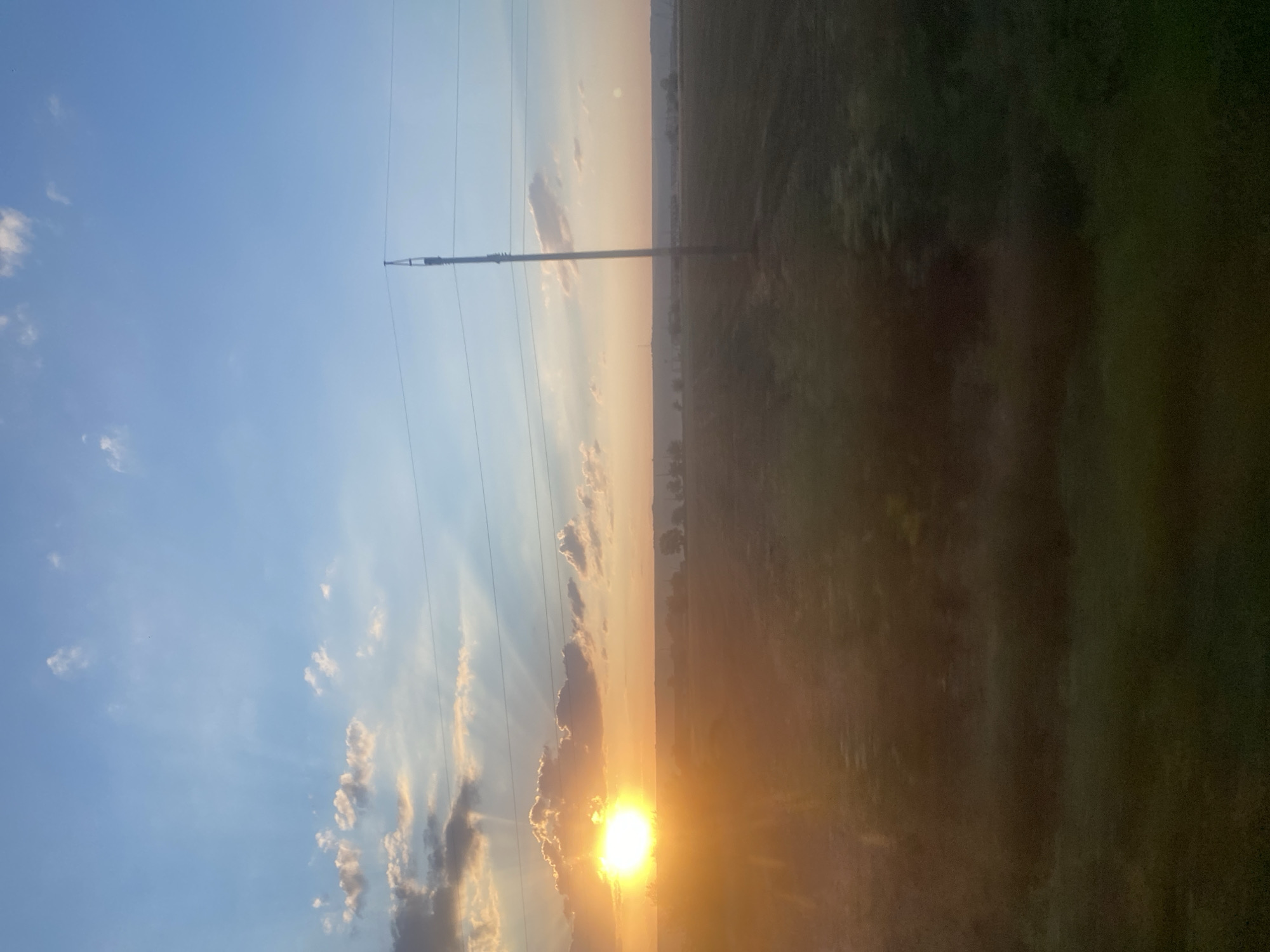 The image captures a sunset over a rural landscape, likely near Balabanu, Moldova. The sun, a bright orb, is positioned low on the horizon, casting golden rays through scattered clouds. The sky is a gradient of blue and orange, transitioning to a soft pinkish hue. In the foreground, a dark, textured field stretches out, appearing to be a cultivated crop. A tall utility pole with power lines is a prominent vertical element on the right side of the frame, silhouetted against the colorful sky. In the distance, a hazy line of trees and structures suggests a small settlement or agricultural buildings. The overall impression is one of natural beauty and a serene rural evening.