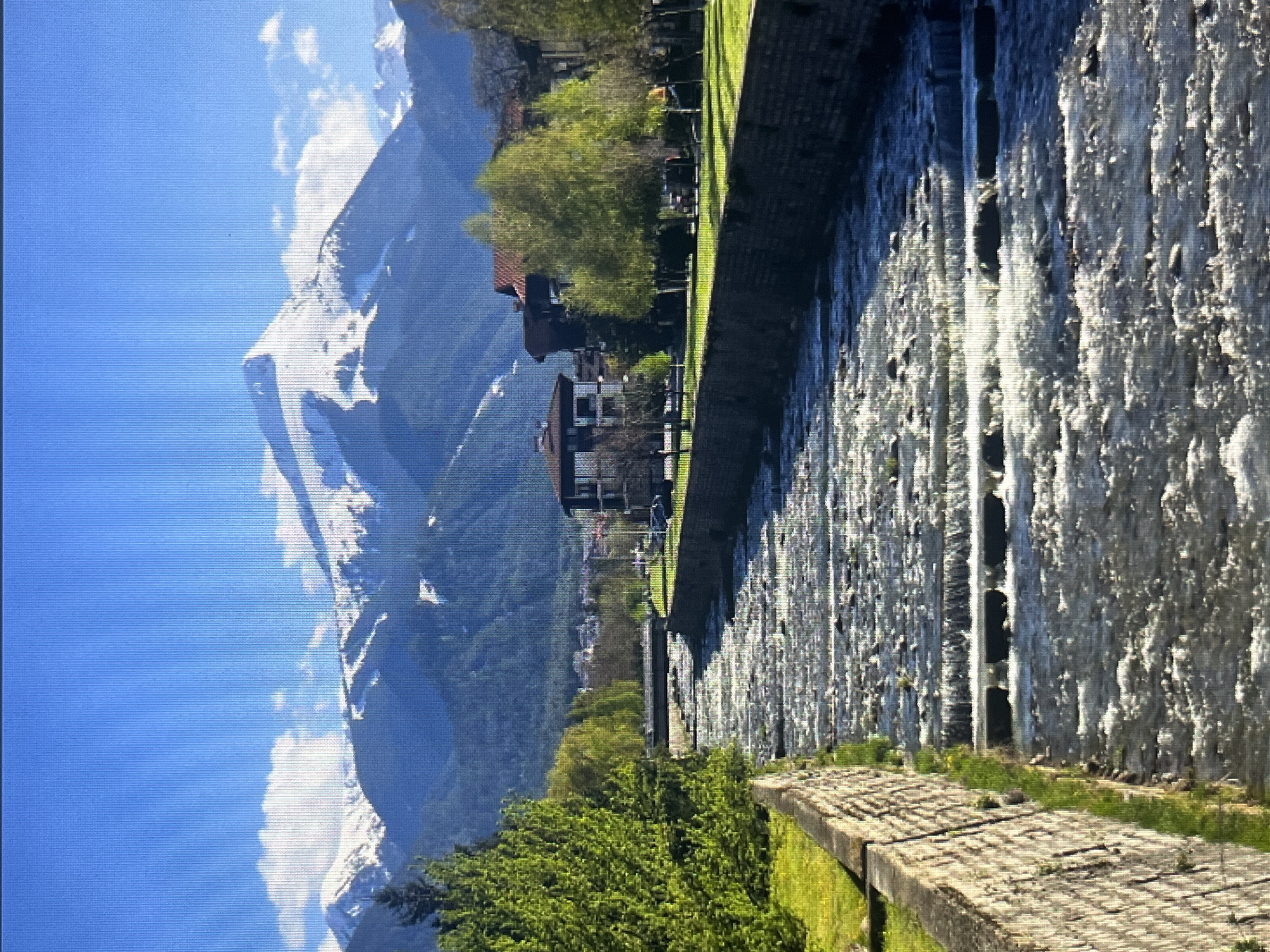 A wide, fast-flowing river with multiple small cascades runs through the center of the image. The riverbanks are lined with stone walls and lush green grass. Beyond the river, there are several buildings with red-tiled roofs and modern architecture, surrounded by trees. In the background, a majestic mountain range with snow-capped peaks dominates the horizon under a clear blue sky with scattered white clouds. The scene appears to be a scenic view of a town nestled in a valley with mountains in the distance. The image does not provide any specific location information within Varna, Bulgaria, nor does it depict any people or ongoing actions.