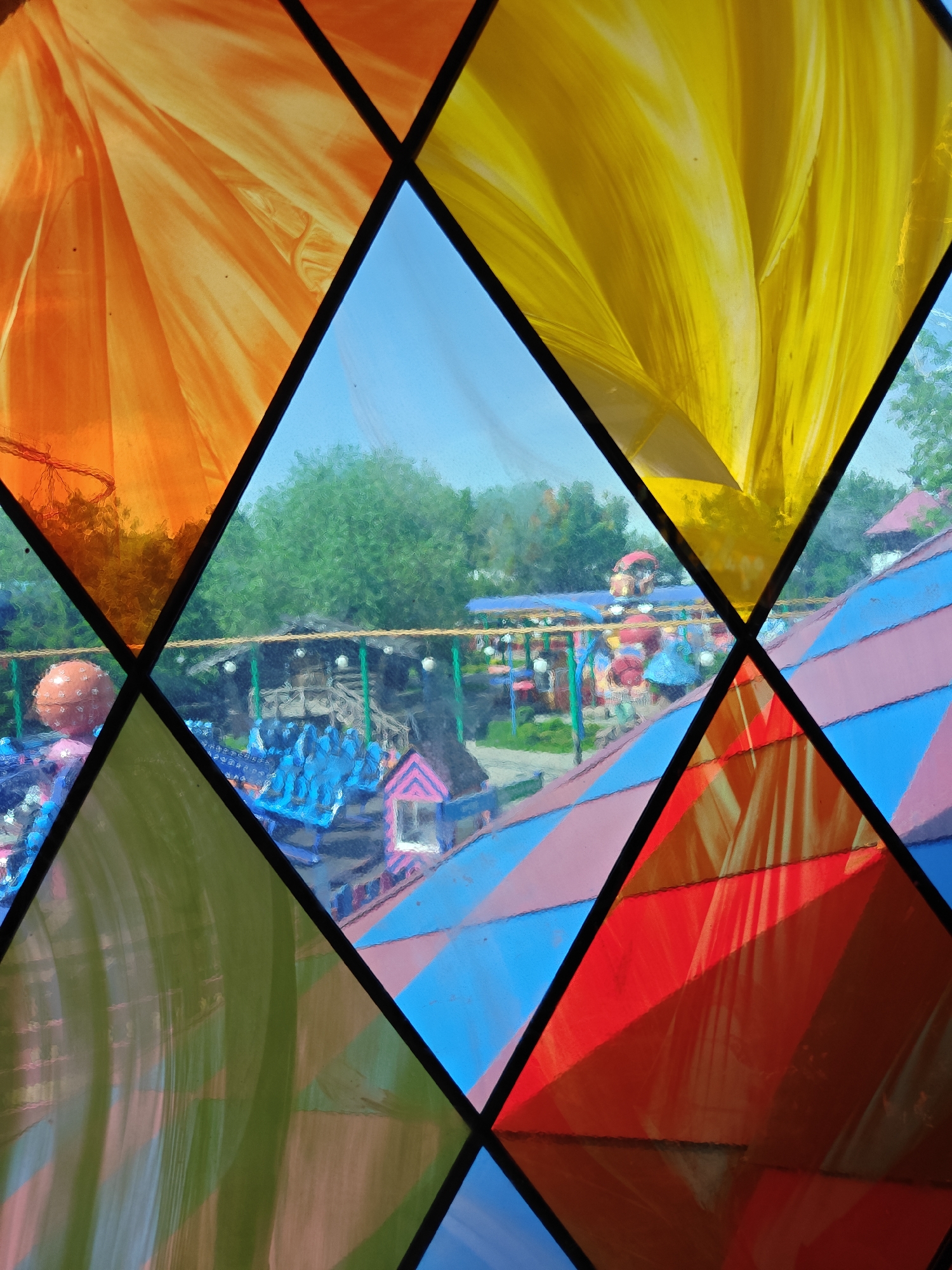 A colorful, close-up view through a diamond-patterned stained glass window offers a distorted glimpse of an amusement park in Khimki, Russia. The window's panes are filled with vibrant hues: orange, yellow, blue, green, and red, each having a swirling, translucent texture. Beyond the stained glass, an outdoor amusement park scene unfolds. Blue-clad figures stand under a striped, multi-colored structure with a pink roof. A row of hanging lights illuminates a rough, wooden fence. In the background, dense green trees and a clear blue sky frame the scene. The image is shot from a slightly low angle, emphasizing the geometric patterns of the window.
