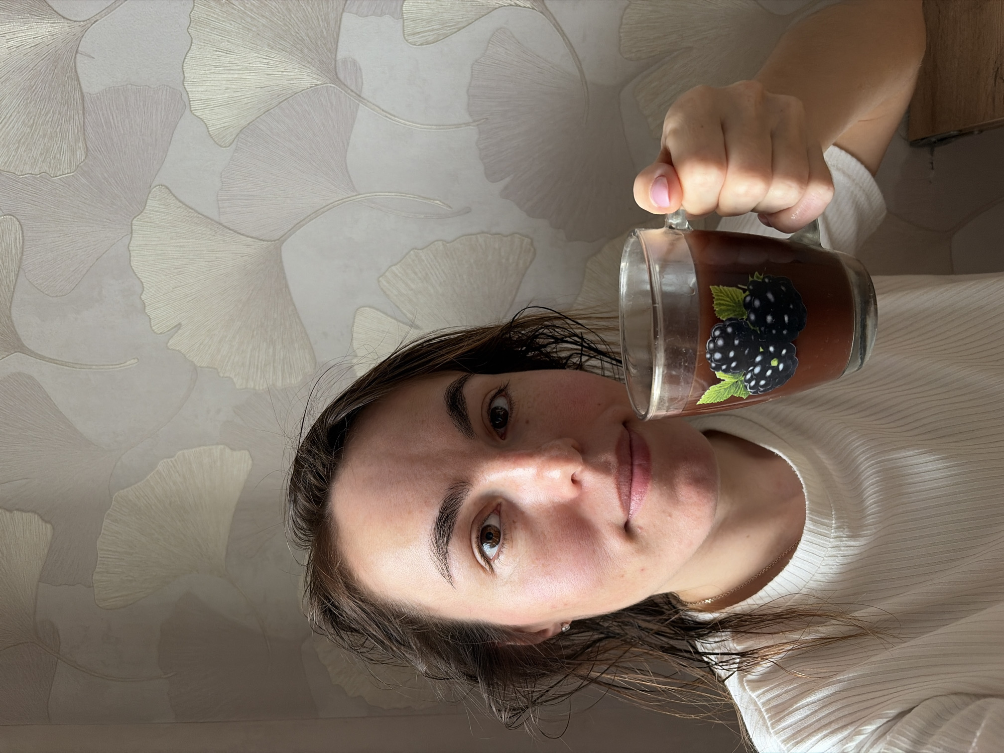 A woman with brown hair and eyes holds a glass mug with a blackberry design, filled with a brown liquid, possibly hot chocolate. She wears a white, ribbed long-sleeved shirt and a delicate gold necklace. Her skin is fair with freckles. The background is a wall with a ginkgo leaf pattern in shades of white and beige. The photo's perspective is slightly skewed, as if taken from below. The location is not determinable to be Izhevsk, Russia based on visual clues alone.