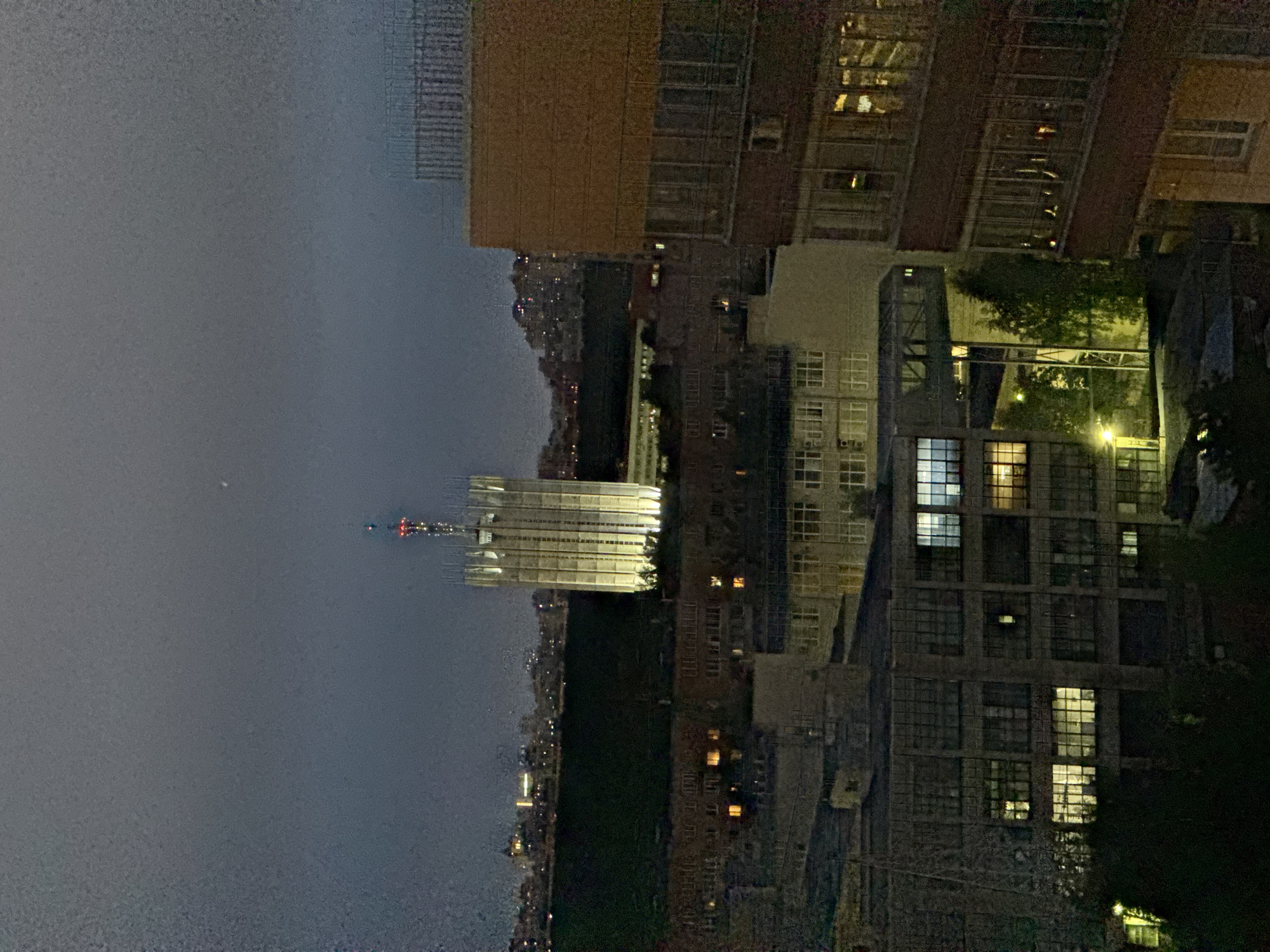 The image depicts a night scene of an urban area, likely in Unknown, Russia. In the foreground are residential and commercial buildings, with many windows illuminated. The building on the left is under construction, its scaffolding and framework visible. In the background, a cityscape stretches into the distance, fading into the dark sky. Patches of green space are also visible, possibly parks or gardens.