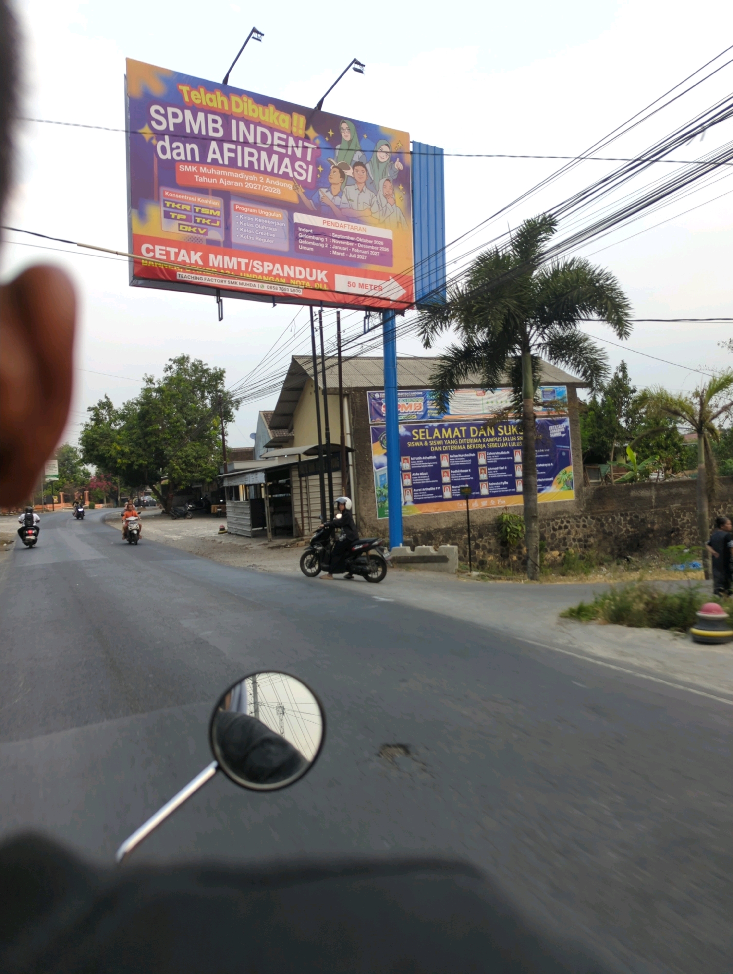 The image, taken from a motorcycle in motion, captures a street scene in Kacangan, Indonesia. On the left, a large billboard announces "SPMB INDENT dan AFIRMASI" for SMK Muhammadiyah 2 Andong, indicating admissions are open for the 2027/2028 academic year. It lists program unggulan (leading programs) such as "Kelas Keberkarian," "Kelas Olahraga," "Kelas Creative," and "Kelas Regular," along with "Konsentrasi Keahlian" (area of expertise) including TKR, TS, TP, TKJ, and DKV. The billboard also provides registration details.
Further down the street, another billboard reads "SELAMAT DAN SUKSES SISWA & SISWI YANG DITERIMA KAMPUS JALUR INDENT DAN DITERIMA BEKERJA SEBELUM LULUS." (Congratulations and success to students accepted through the indent campus route and accepted for work before graduation.) This billboard displays names and faces of individuals.
Several motorcycles are visible on the road. On the right, a person is riding a motorcycle with a helmet. In the background, buildings with tiled roofs and a stone wall are present, along with trees, including a palm tree. Electrical wires are strung overhead. The road surface appears to be asphalt with some cracks. The overall lighting suggests it is daytime, possibly late afternoon due to the soft light.