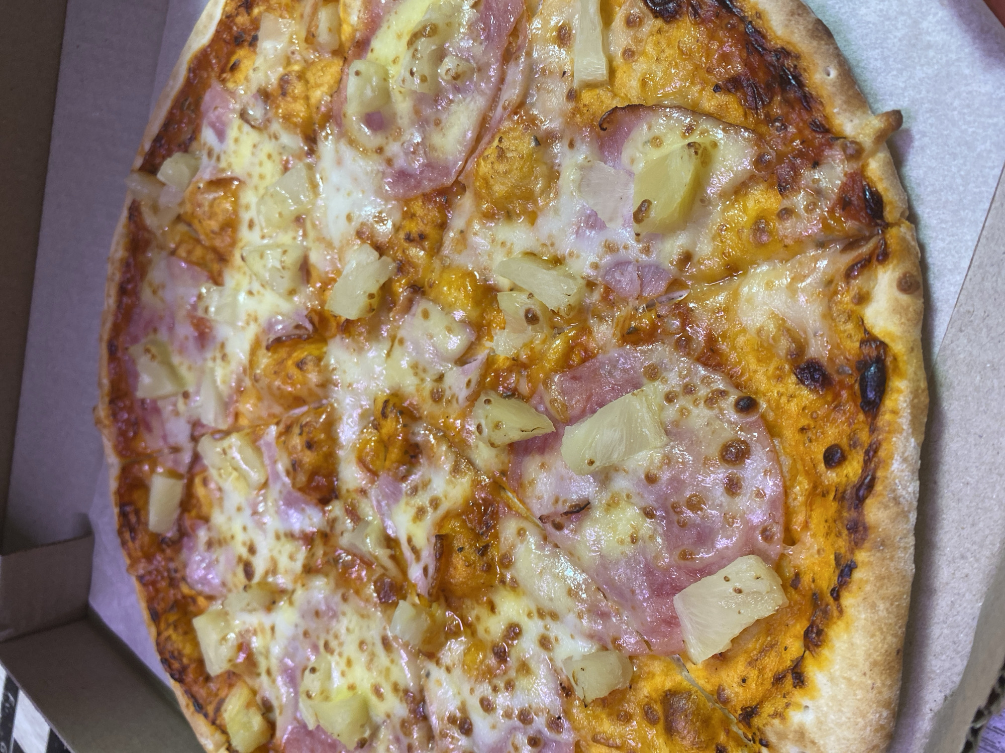 This image displays a close-up, overhead view of a Hawaiian pizza housed within a cardboard pizza box. The pizza is generously topped with melted, bubbly white and light yellow cheese, numerous pale yellow, irregularly cut pineapple chunks, and slices of light pink ham. The cheese exhibits areas of golden-brown caramelization, and the reddish-orange tomato sauce base is visible between the toppings. The pizza's crust is golden-brown with darker, slightly charred edges and some browned spots, indicating a well-baked finish. The pizza has been pre-sliced into multiple portions, with clear divisions visible. It rests on a white, possibly grease-proof, liner that covers the bottom of the light brown, unbleached cardboard box. The top-left corner and a portion of the left inner wall of the pizza box are visible, showing the textured cardboard material. No people, specific actions, or interactions are depicted. The environment is limited to the interior of the pizza box. The location of Chaikovsky, Russia, cannot be determined from the visual information provided in the image.