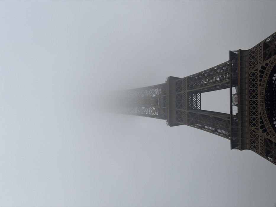 The image displays the Eiffel Tower in Paris, France, enveloped in dense fog. The lower and middle sections of the tower's iron lattice structure are clearly visible, rising from the bottom of the frame. The first and second platforms, along with their intricate support beams and cross-bracing, are distinguishable. The metallic framework exhibits a dark grey hue. As the tower ascends, its upper section gradually fades into and is ultimately obscured by a thick, uniform light grey fog that dominates the upper two-thirds of the sky. The overall lighting is soft and diffused, characteristic of an overcast or heavily foggy day. No individuals, vehicles, or other distinct objects are present within the immediate scene.