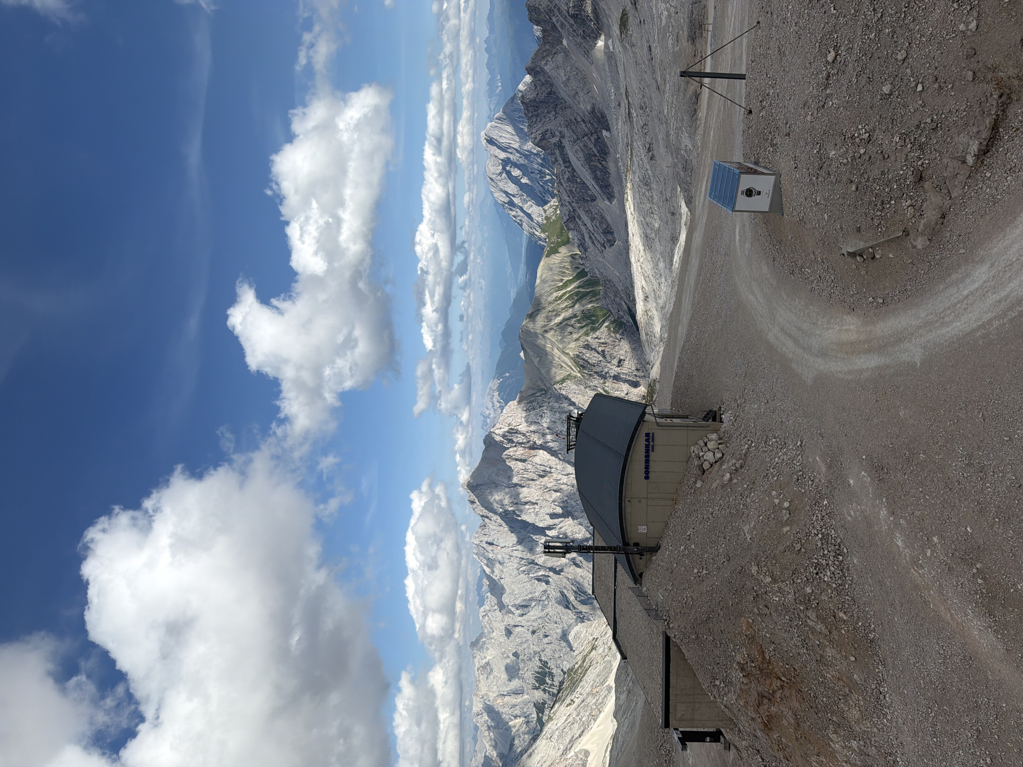 The image shows a mountainous landscape with a building and a dirt road. The building, labeled "SONNENKAR", appears to be a station or a restaurant. The surrounding area is rocky and barren, with snow-capped peaks in the distance. The sky is blue with scattered clouds. There are no people visible in the image. The location is likely the Zugspitze, Germany's highest mountain, which is accessible from Garmisch-Partenkirchen.