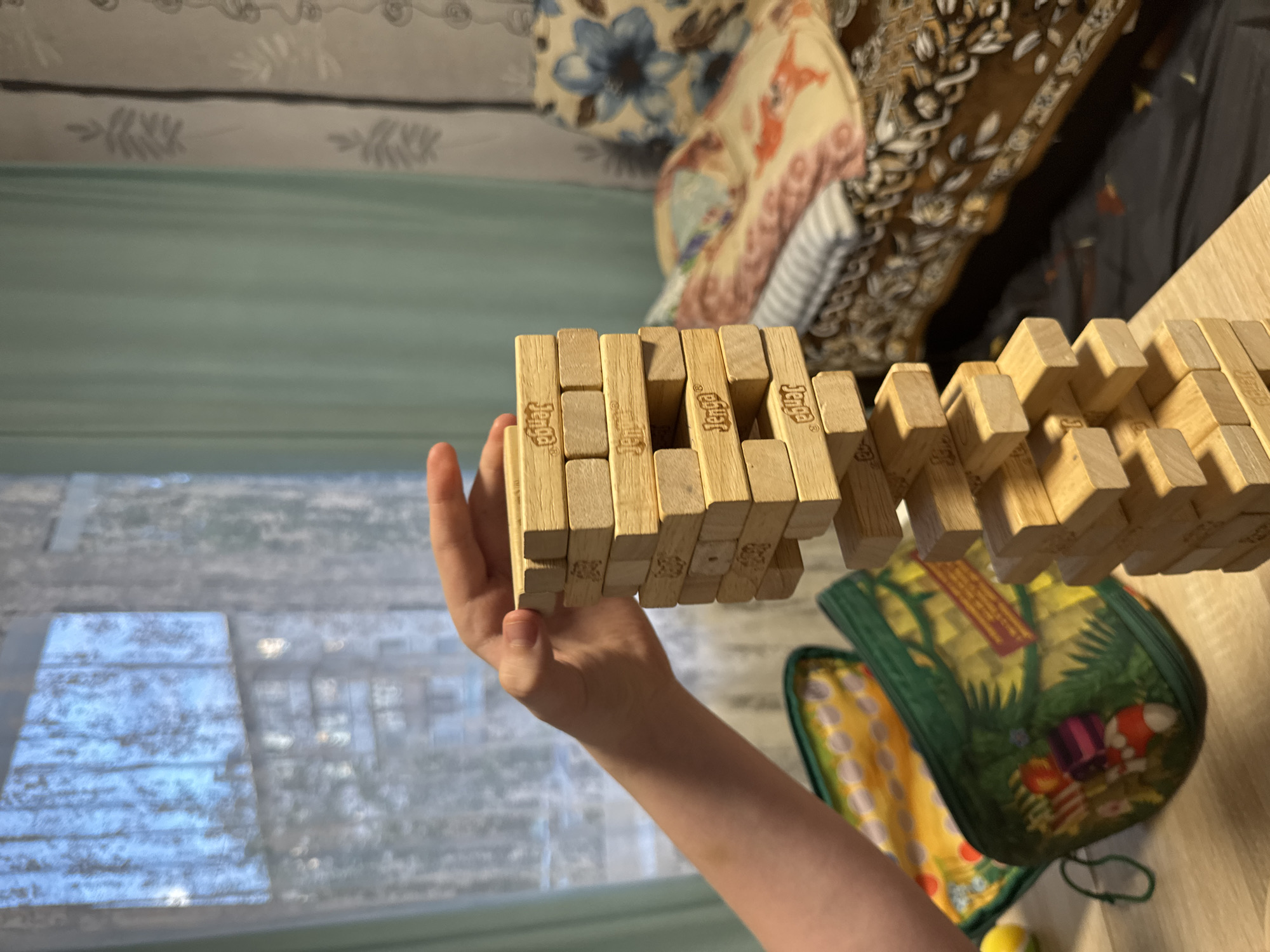 The image depicts a close-up view of a hand attempting to remove a wooden block from a precarious Jenga tower. The Jenga blocks are light brown wood and are stacked in an alternating pattern. The hand is positioned on the right side of the tower, with fingers reaching towards a block. The background reveals a domestic setting. To the left, a green zippered bag with a Christmas-themed design is visible, along with a yellow object that could be a toy or a ball. The background further shows a window with sheer curtains and what appears to be a building outside. On the right, a bed with patterned bedding is visible. The lighting suggests an indoor environment, possibly daytime due to the light coming from the window. The scene is devoid of any specific landmarks or indicators that would place it in Chaikovsky, Russia.