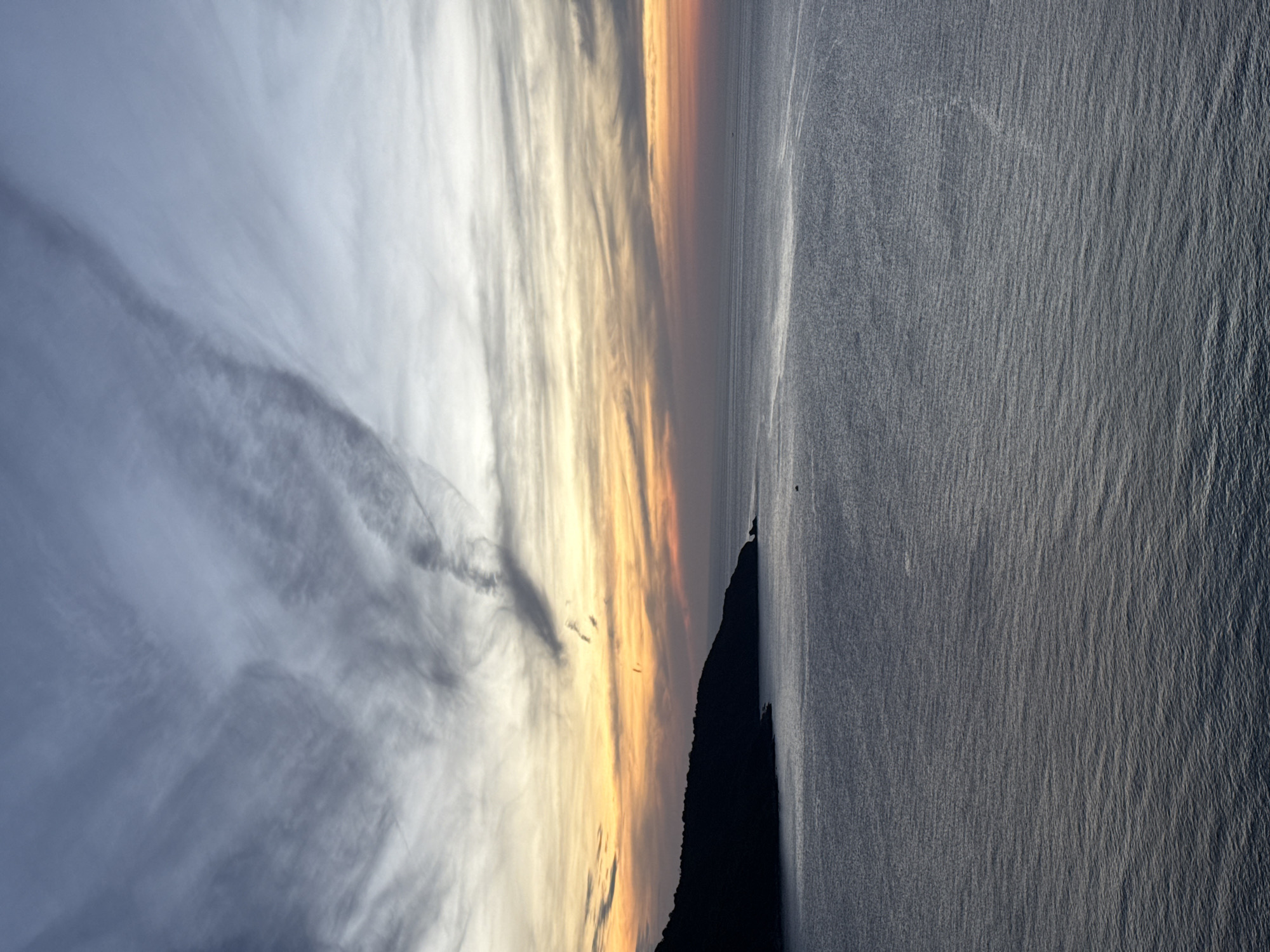 The image depicts a serene seascape at dusk, likely in Da Nang, Vietnam, given the characteristic coastal landscape. The scene is dominated by the vast expanse of the ocean, its surface gently rippling under a twilight sky. The water exhibits a deep, muted blue-gray hue, with subtle variations in tone reflecting the fading light. A dark, silhouetted headland juts out from the left side of the frame, its outline sharp against the softer colors of the sky. The coastline appears rugged and undeveloped, suggesting a natural or minimally impacted environment.
The sky above is a breathtaking canvas of transition. The upper portion is a cool, pale blue, gradually giving way to warmer tones as it approaches the horizon. Streaks of soft orange, yellow, and pink paint a gradient of sunset colors, intermingled with wispy, elongated clouds that add texture and depth. A few faint, elongated contrails are visible in the sky, likely from aircraft.
In the distance, a small, dark object is visible on the water, possibly a boat or a buoy, adding a sense of scale to the immense seascape. The overall atmosphere is tranquil and contemplative, capturing the ephemeral beauty of the day's end by the sea. The absence of any discernable human figures or structures contributes to the natural and undisturbed feel of the landscape.