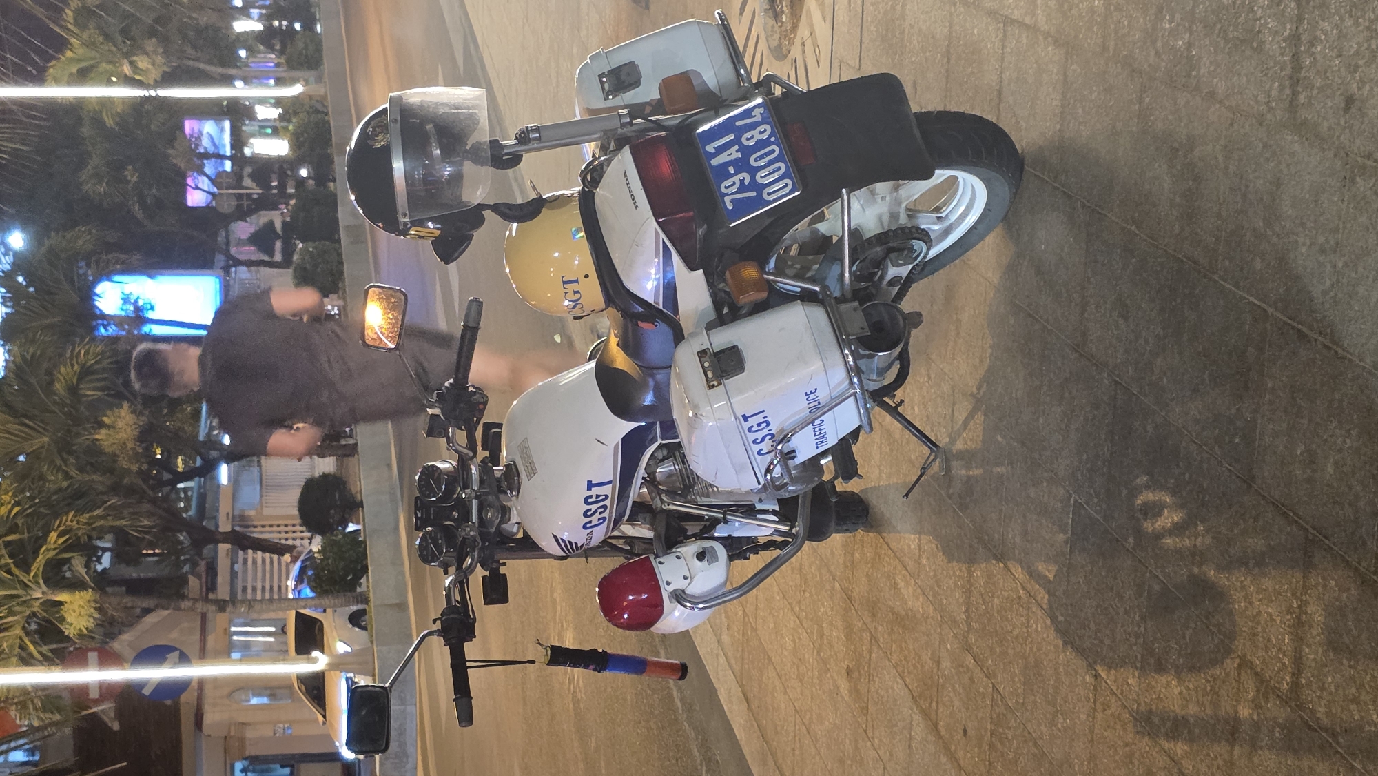 A high-angle photograph depicts a white Honda motorcycle, identified as traffic police ("C.S.G.T. Traffic Police"), parked on a tiled surface in Nha Trang Ward, Vietnam. The license plate number is "79-A1 78000". The motorcycle is equipped with police accessories, including storage compartments and a helmet. A person wearing a dark shirt is seen standing behind the motorcycle. The background features urban elements, including streetlights, palm trees, and buildings.