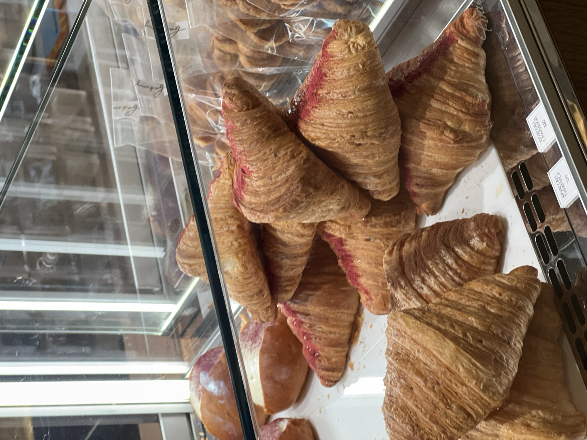 The image shows a close-up of croissants in a display case, likely in a bakery or cafe. Some croissants have a pink topping or coating on the ends. The scene appears to be inside a business establishment. Some of the croissants are labeled with tags that have text in Russian, and the location is Vladivostok, Russia. The focus is on the pastries in the display.