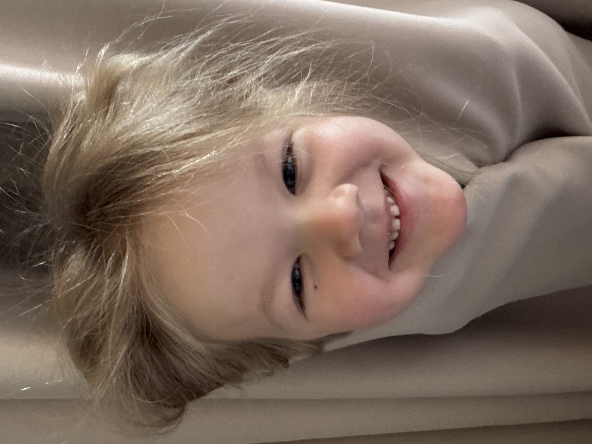 A close-up, angled shot depicts a young child with fair skin and blonde hair. The child is smiling, revealing a set of baby teeth. A mole is visible on the child's cheek, close to the eye. The child appears to be partially covered or wrapped in a beige or light tan fabric. No identifiable location is discernible from the image, so its relationship to Ферма, Russia, is unknown.