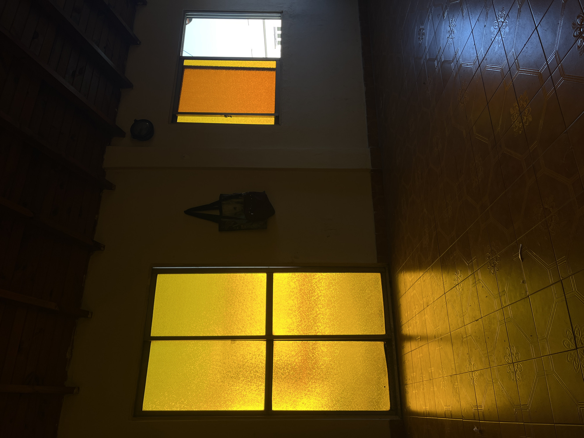 This image, taken in Buenos Aires, Argentina, depicts an interior space, possibly a stairwell or a room with high ceilings, featuring two prominent windows. The original orientation appears to be rotated 90 degrees clockwise, placing a tiled floor on the right and a wooden ceiling/upper wall structure on the left.
The upper window consists of two panes: a clear upper pane revealing bright daylight outside and a lower pane of translucent, textured orange-tinted glass. Below it, a larger window is divided into four sections, all fitted with translucent, textured yellow-tinted glass, through which sunlight casts a warm glow. The walls are a light, possibly cream or pale yellow, color.
On the left side of the frame, a dark, sloped wooden structure with visible beams suggests a ceiling or an upper architectural element. On the right, the floor is covered with brownish-orange tiles, embossed with a floral or geometric pattern. A striking rectangular patch of intense yellow light from the sun streams through one of the lower windows, illuminating a section of the tiled floor and casting sharp shadows.
Between the two windows, an angular, dark decorative object is mounted on the wall. Above the upper window, a small, dark circular object, possibly a clock, is faintly visible. The strong, direct sunlight and the colored light filtering through the windows indicate it is daytime, likely morning or late afternoon. The scene is static, with no activity taking place, and no visible text is present.