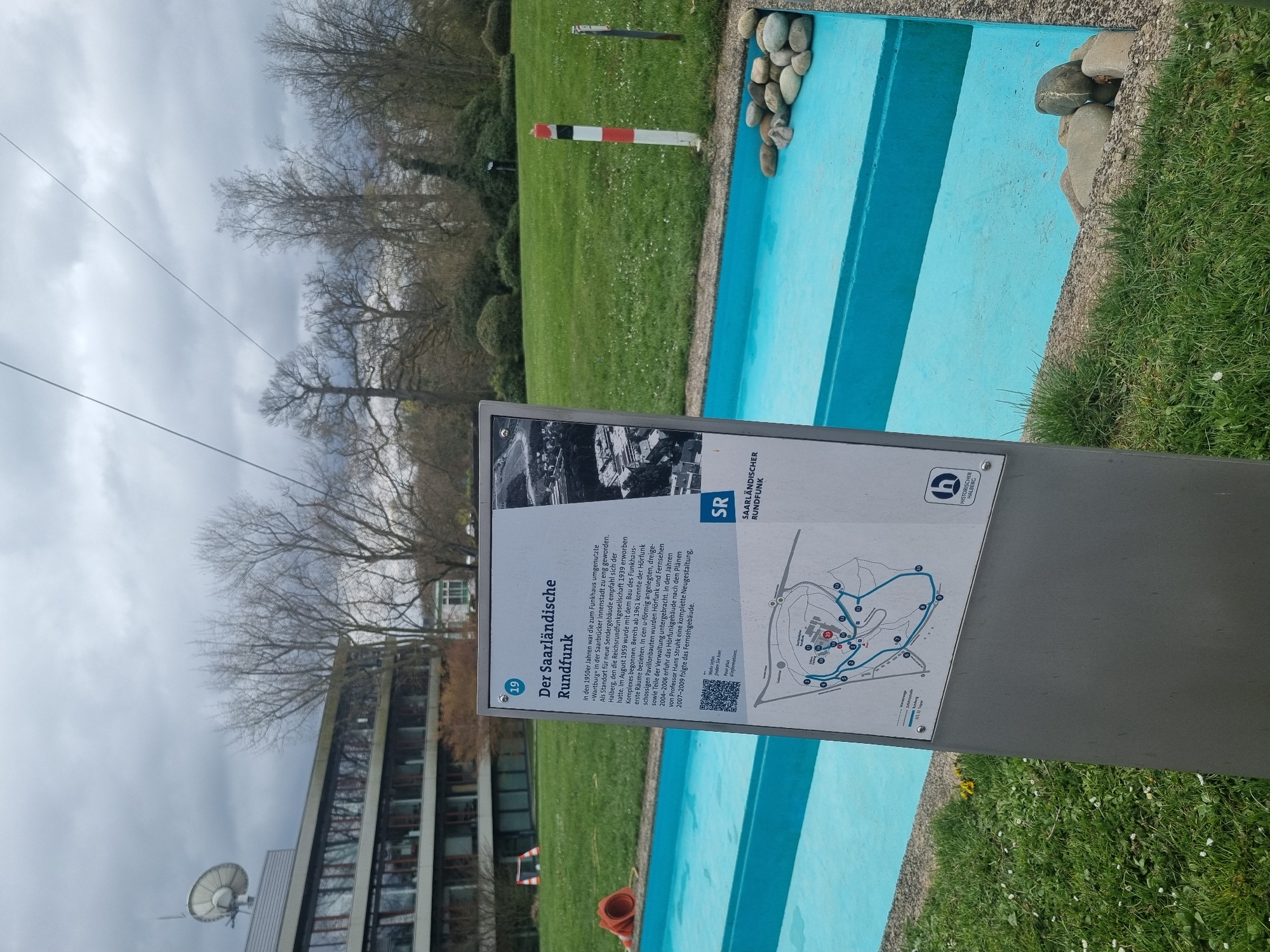 A photograph taken in Saarbrücken, Germany, depicts a sign on the Halberg detailing the history of the Saarländischer Rundfunk (SR), the Saarland state broadcasting corporation. The sign provides information about the radio station and includes a QR code, a map of the area, and the SR logo. The image also shows a turquoise colored, tiered water feature with stone edging. The surrounding environment includes a building with a satellite dish, trees, grass, and a red-and-white pole. The sky is overcast.