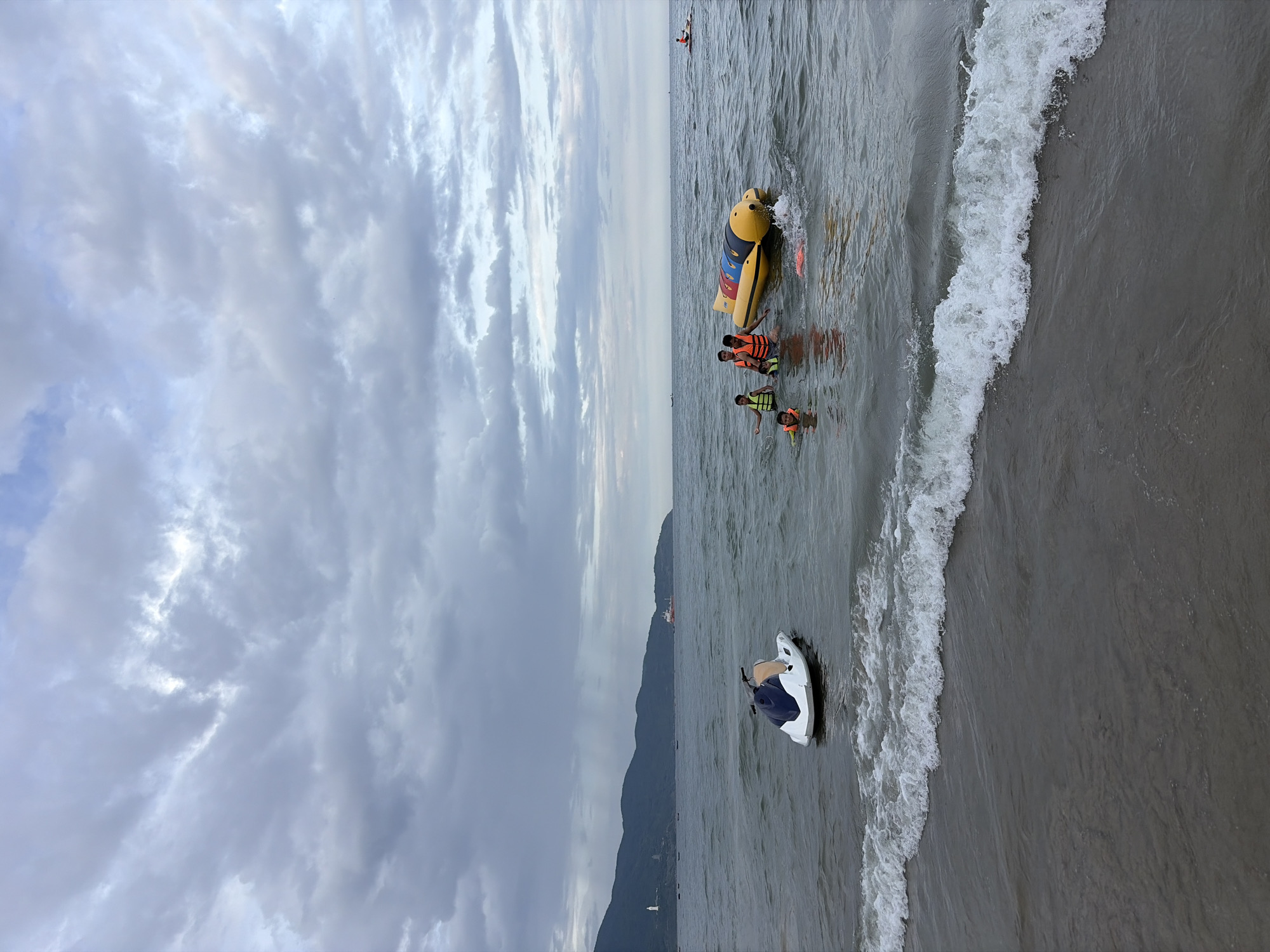The image depicts a group of people enjoying recreational activities on the ocean at Mỹ Khê Beach in Đà Nẵng, Vietnam. The weather appears overcast with dramatic clouds in the sky. In the foreground, a jet ski is partially submerged near the shore. Further out in the water, a group of individuals, including children wearing life vests, are interacting with a yellow inflatable banana boat. One person is seated on the banana boat, while others are in the water around it, some appearing to be holding onto it. In the middle ground, a larger boat with a red structure is visible in the distance, near the coastline which is lined with mountains. Another individual is seen on a personal watercraft further to the right. The waves are gently rolling onto the sandy shore.