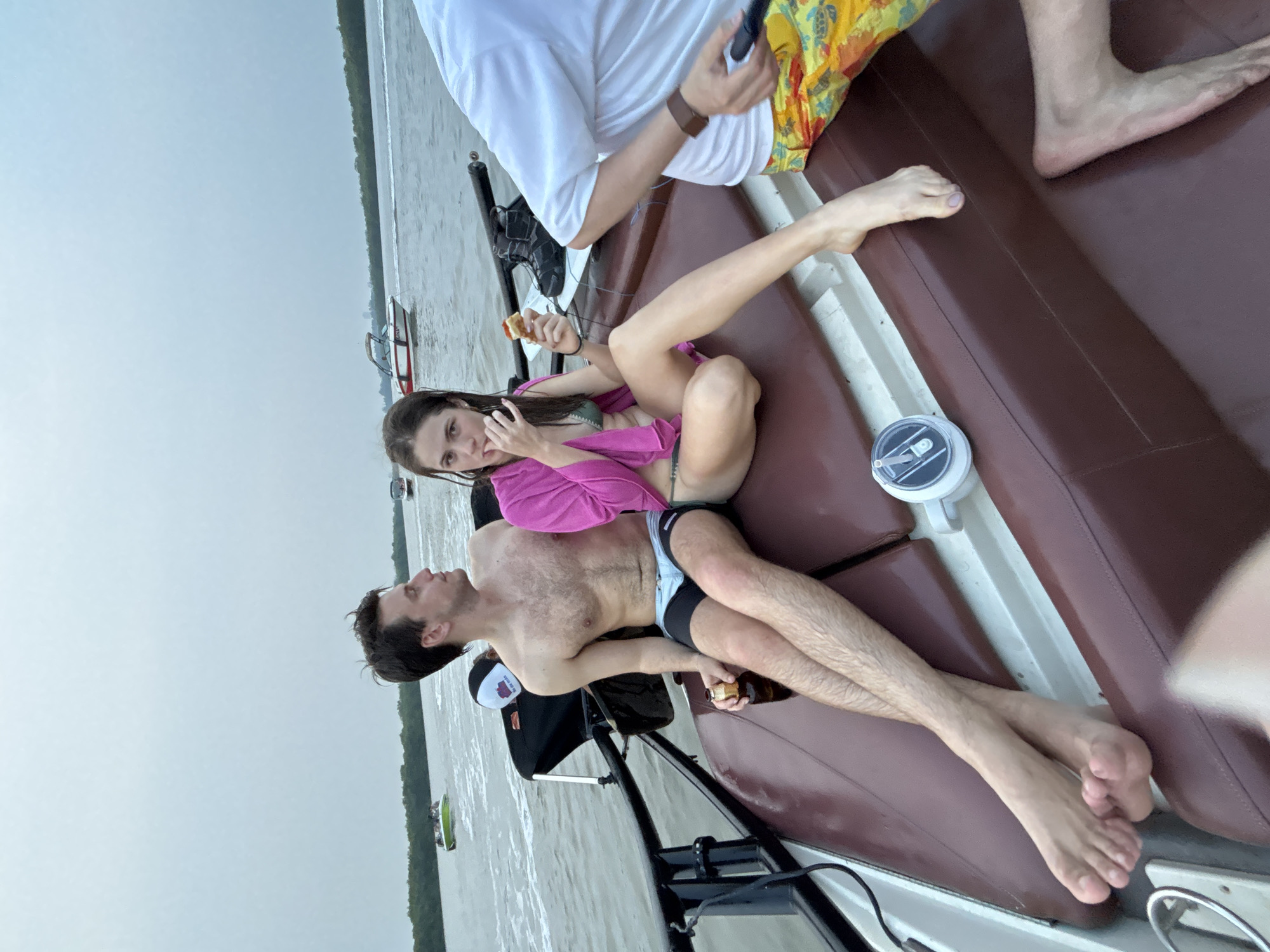A slightly rotated medium shot depicts four individuals on a boat in Unknown, Unknown. A woman with long dark hair, wearing a green bikini and a pink cover-up, sits and eats pizza. A man with fair skin and dark hair lies on the seat next to her, shirtless and wearing black and blue swim trunks. He is holding a bottle. Another individual is partially visible, wearing a white shirt and colorful shorts, holding a phone. Bare feet of a fourth person are visible in the upper right corner of the image. The boat is on the water, with trees visible on the shoreline. The sky is overcast.